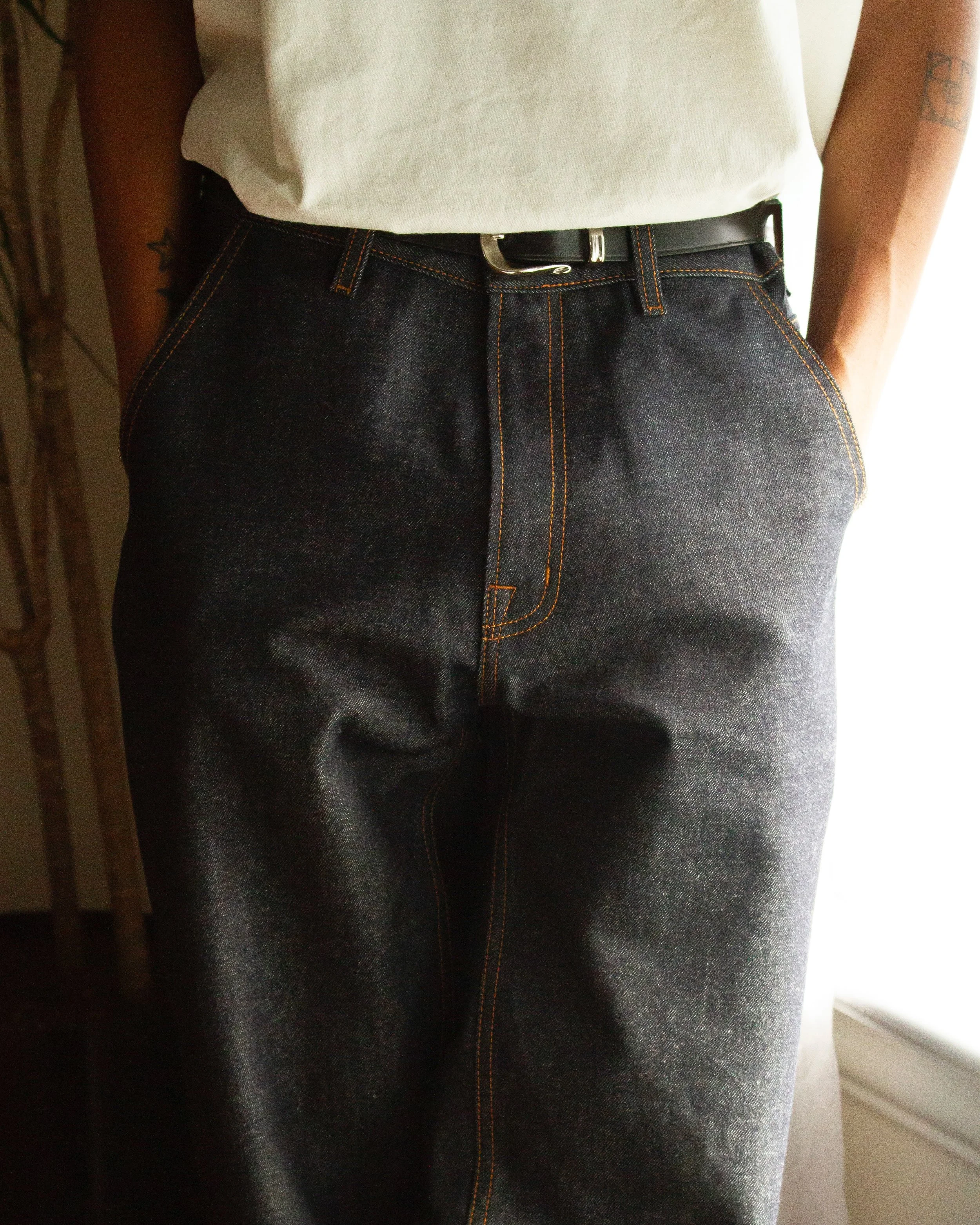 Close-up of a person wearing dark denim jeans with orange stitching, a white t-shirt, and a black belt with a silver buckle, standing indoors near a window.