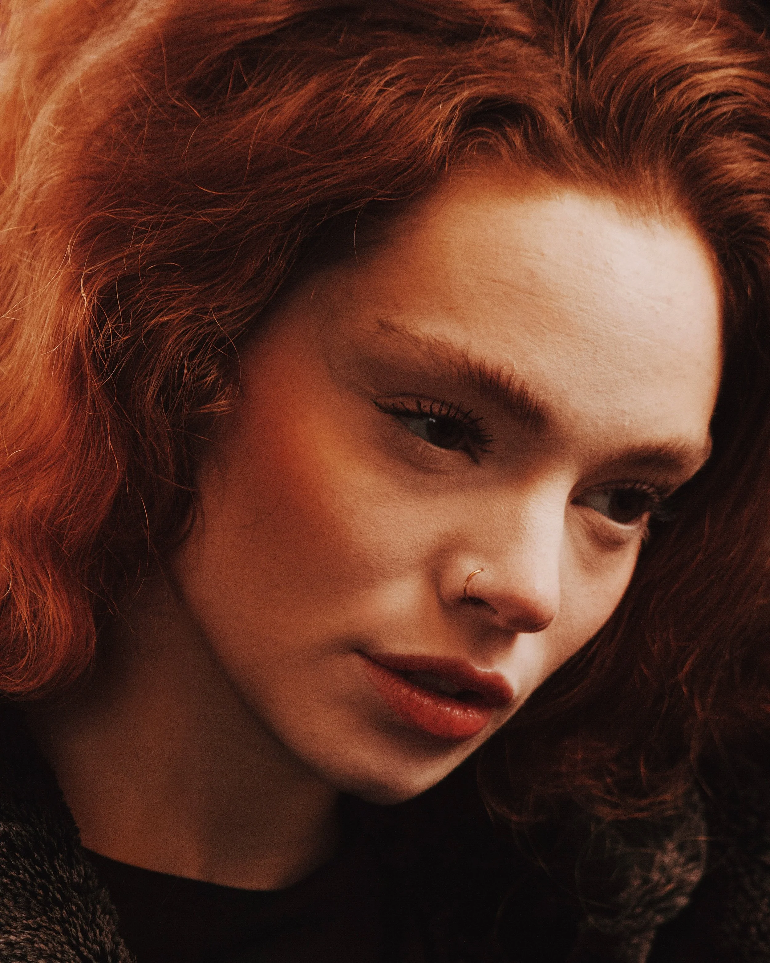 Close-up of a young woman with red hair, wearing a nose ring, looking down thoughtfully.