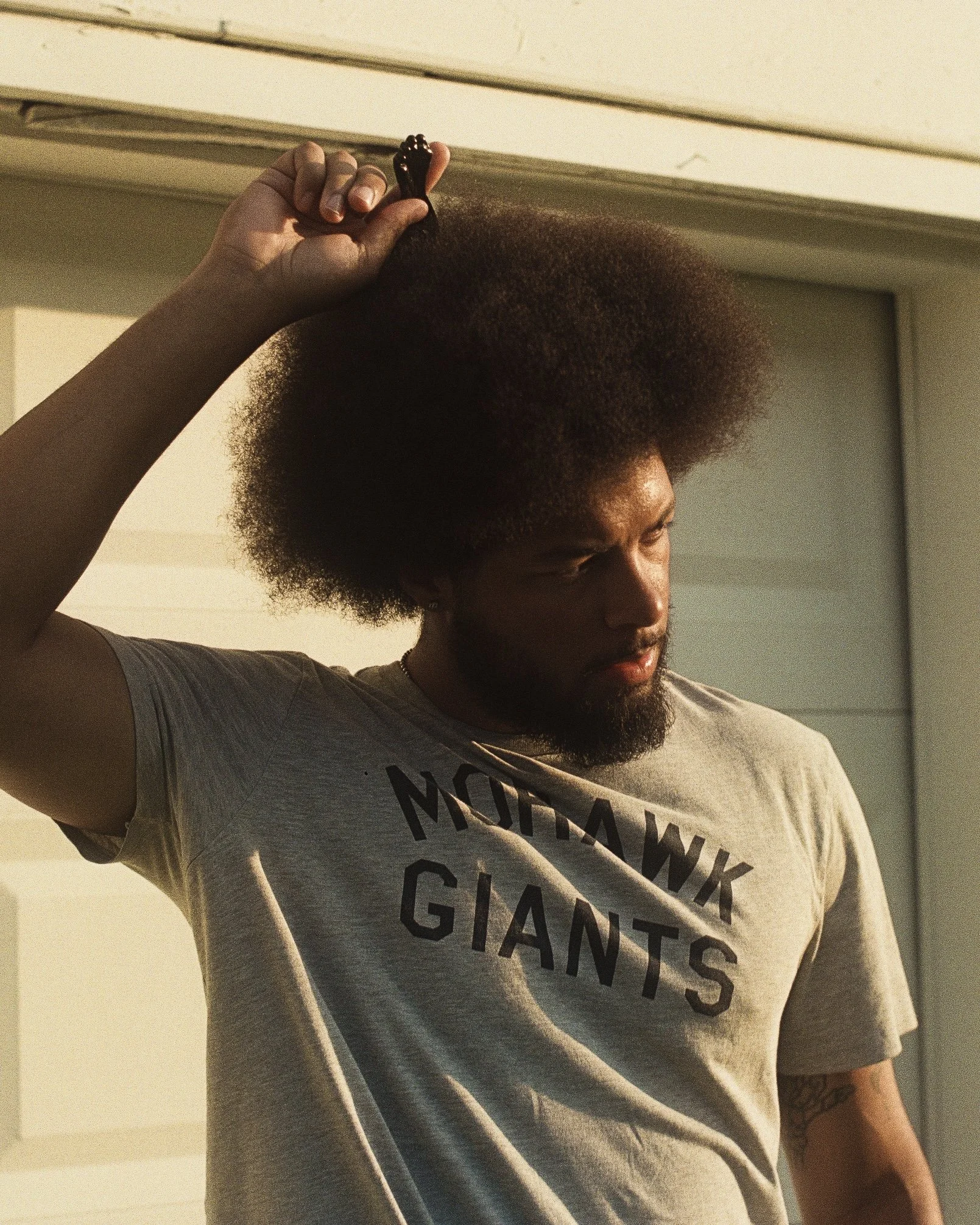 A man with a large afro hairstyle, wearing a gray T-shirt, adjusting his hair with his right hand.
