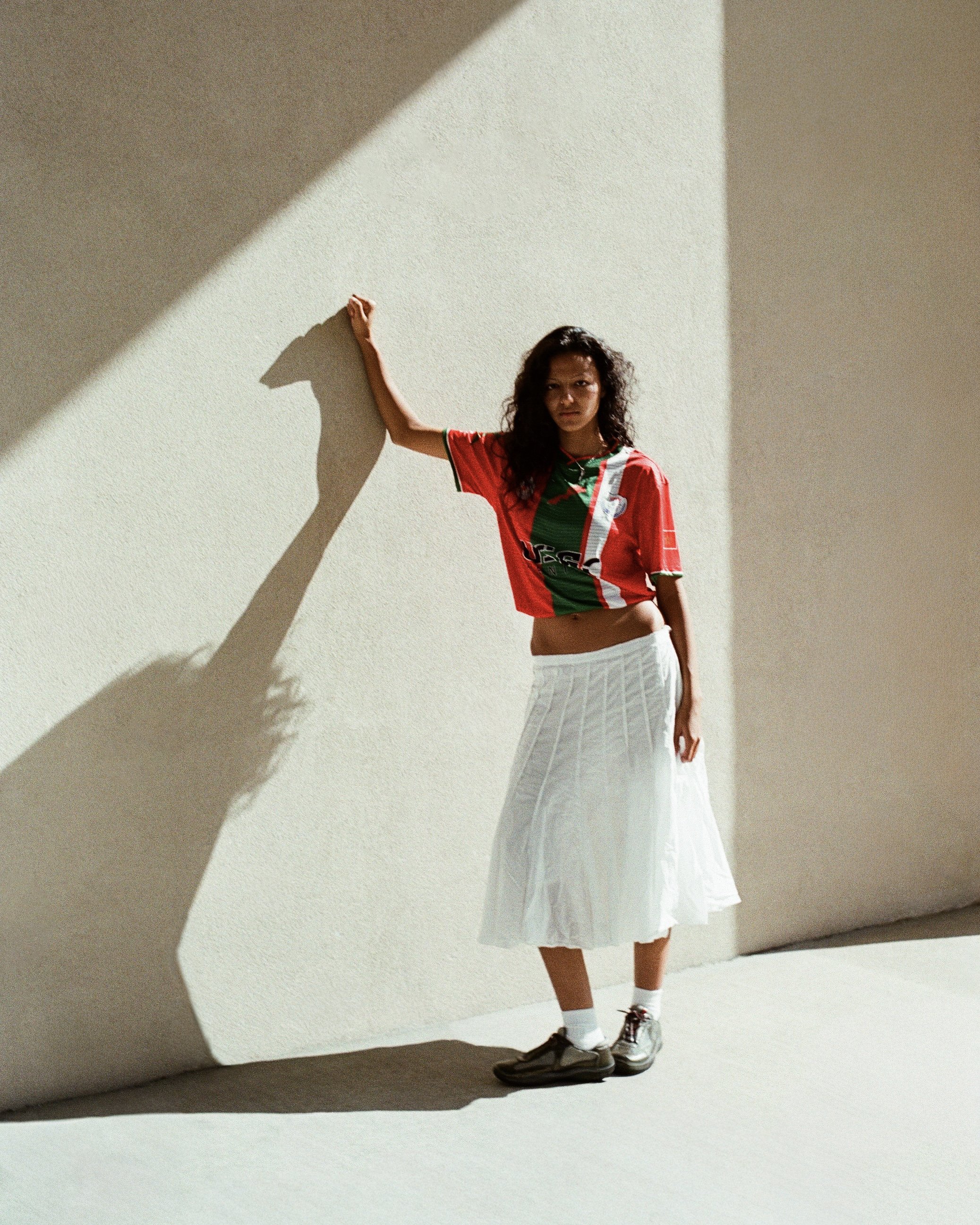 Woman standing against a beige wall, casting a shadow. She wears a red and green sports jersey, a white pleated skirt, white socks, and sneakers.