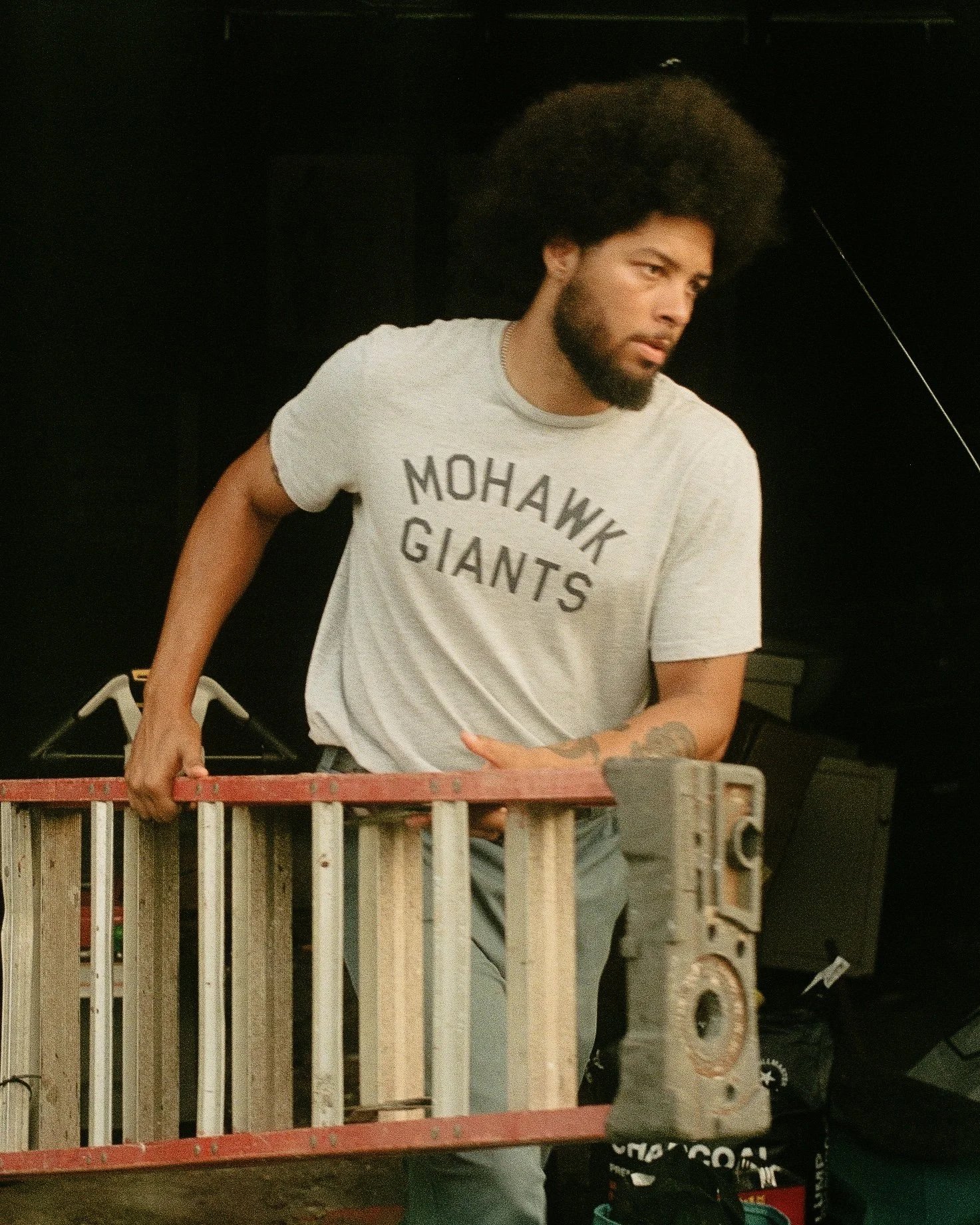 A man with curly hair, beard, and tattoos on his arm, wearing a Mohawk Giants t-shirt, is standing behind a ladder and looking to his right in a dark room.