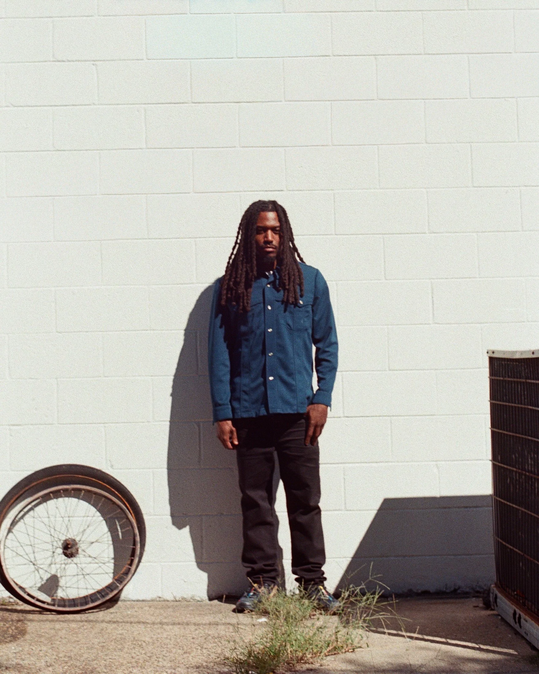 A man standing against a white brick wall with long dreadlocks, wearing a blue jacket, black pants, and sneakers. An old bicycle wheel and a metal container are on either side of him.