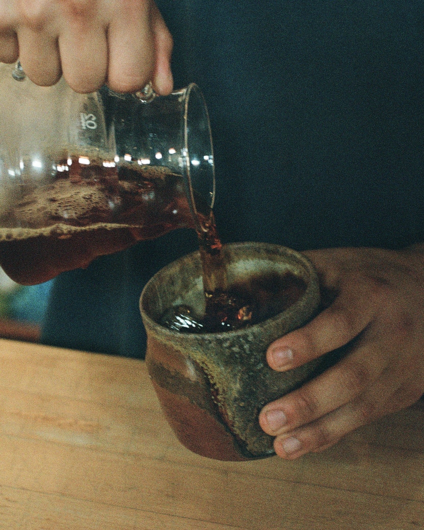 A person pouring a dark beverage from a glass into a rustic ceramic cup, hands visible, on a wooden surface.