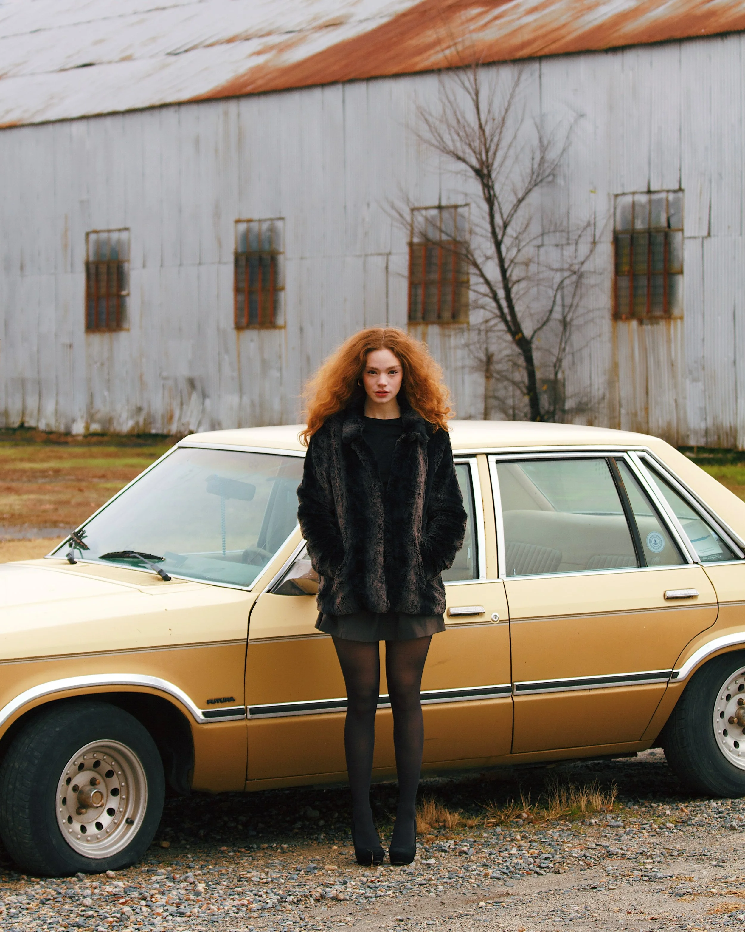 A woman with curly red hair standing next to a beige vintage car in front of a weathered wooden building with a leafless tree behind her.