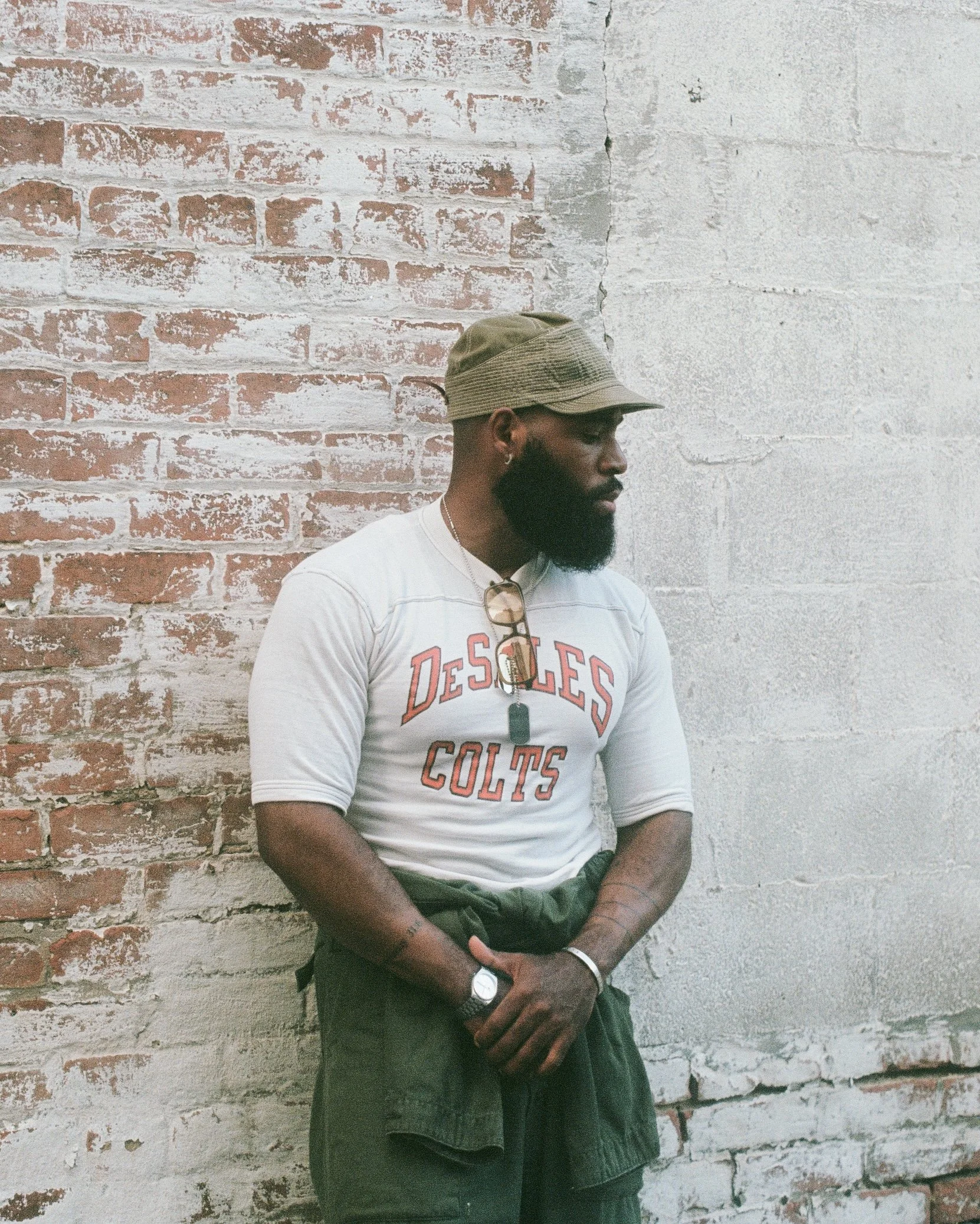 A man with a beard and glasses hanging from his shirt collar is standing against a brick wall and looking to the side, wearing a tan cap, white t-shirt, a watch, and a bracelet.