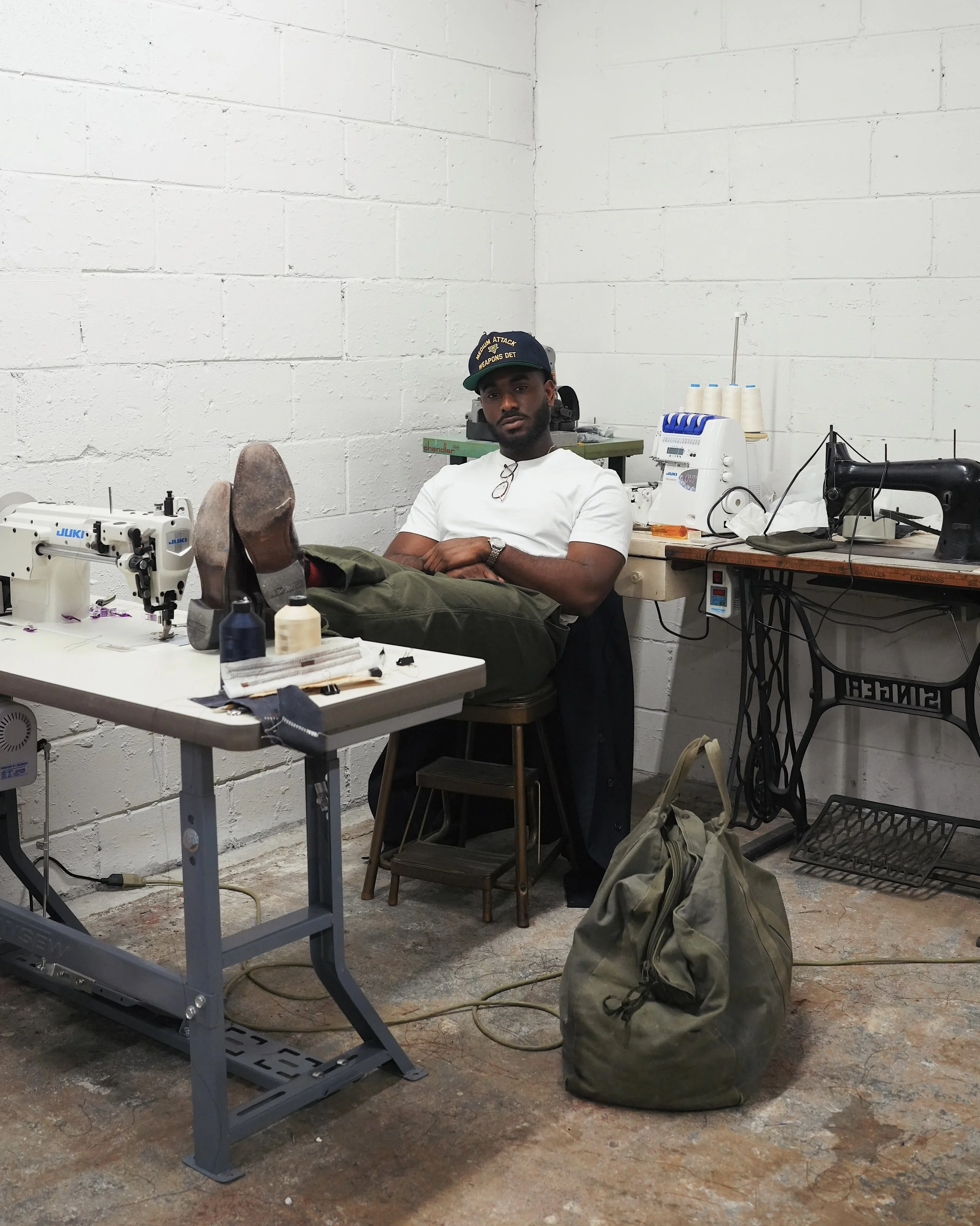 A man sitting with his feet up on a sewing machine table in a workshop, surrounded by sewing equipment and supplies.