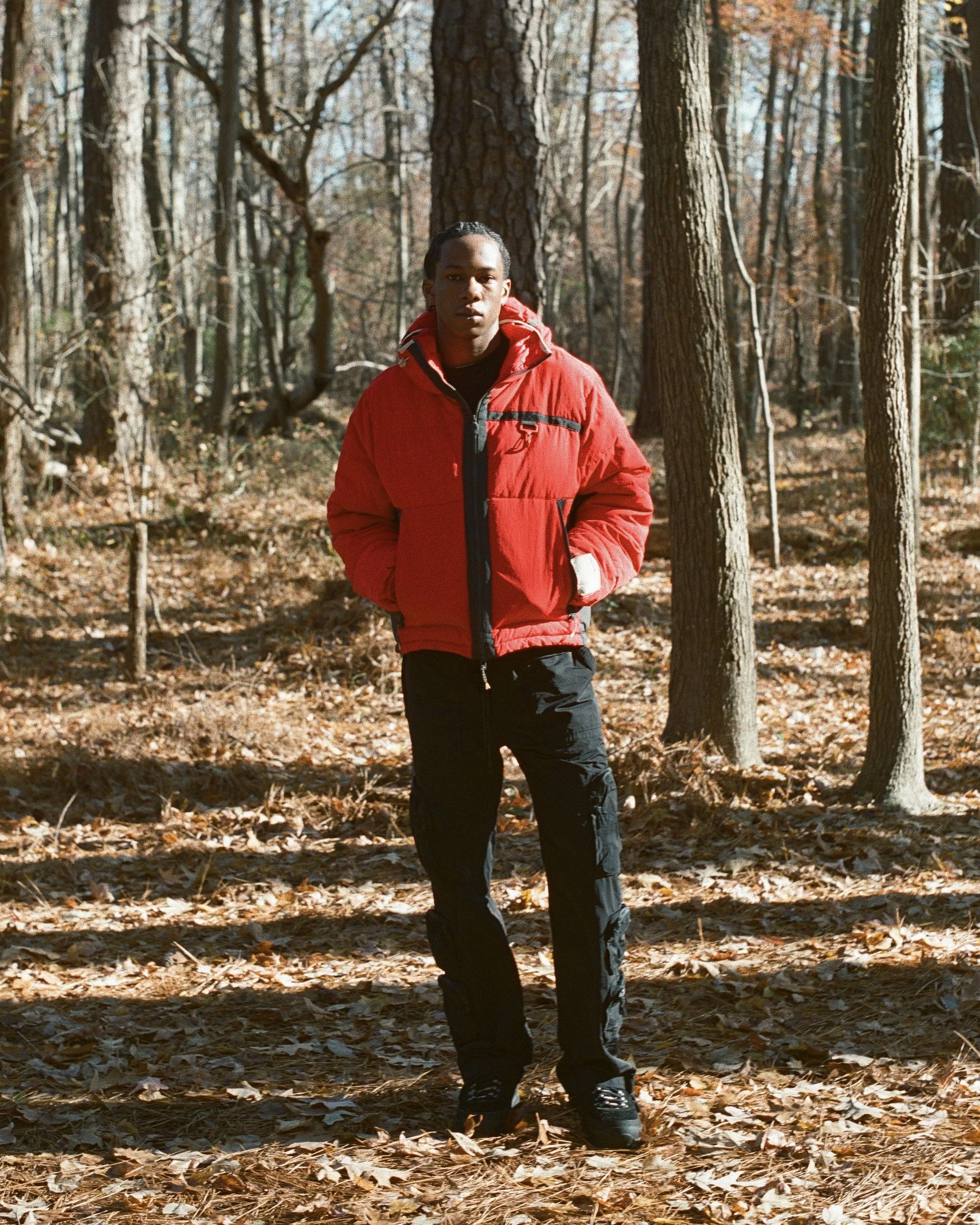 A person standing in a wooded area during fall, wearing a red jacket and black pants.