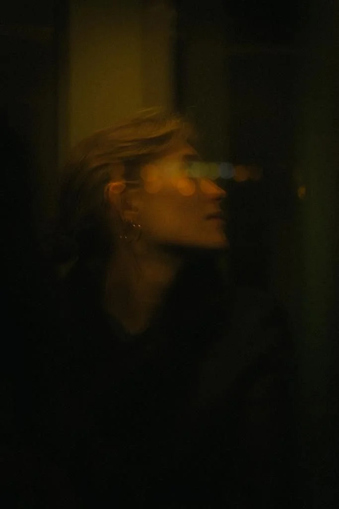 Eine junge Frau mit kurzen, blonden Haaren in einem dunklen Raum, bei Nacht, reflektierende Lichter auf ihrer Brille, der Stil ist künstlerisch und atmosphärisch.