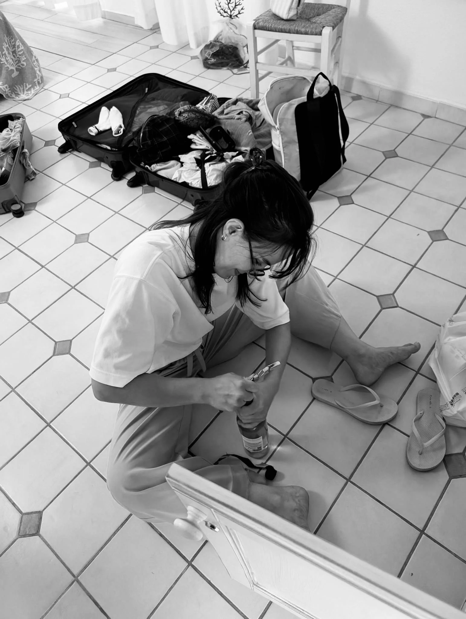 Frau sitzt auf dem Boden, öffnet eine Getränkeflasche, auf dem Boden liegen Flip-Flops, Koffer mit Kleidung und andere Reiseutensilien an ihrer Seite, im Hintergrund ein Stuhl, ein kleiner Tisch und eine Pflanze.