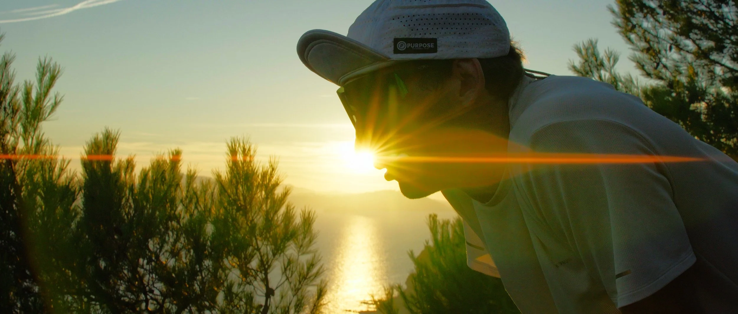 A person wearing sunglasses, a white cap, and a gray shirt standing outdoors during sunset with the sun creating a lens flare, surrounded by trees.