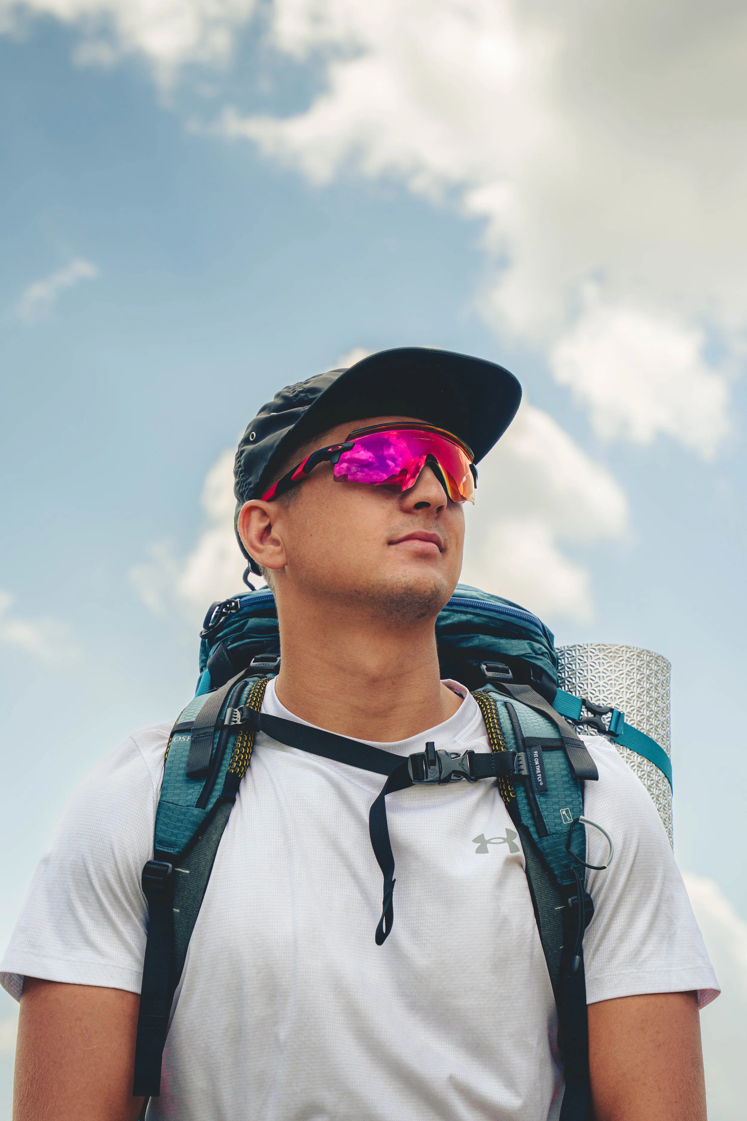 A man wearing a black cap, pink reflective sunglasses, and a white shirt with a backpack, outdoors with a cloudy sky in the background.