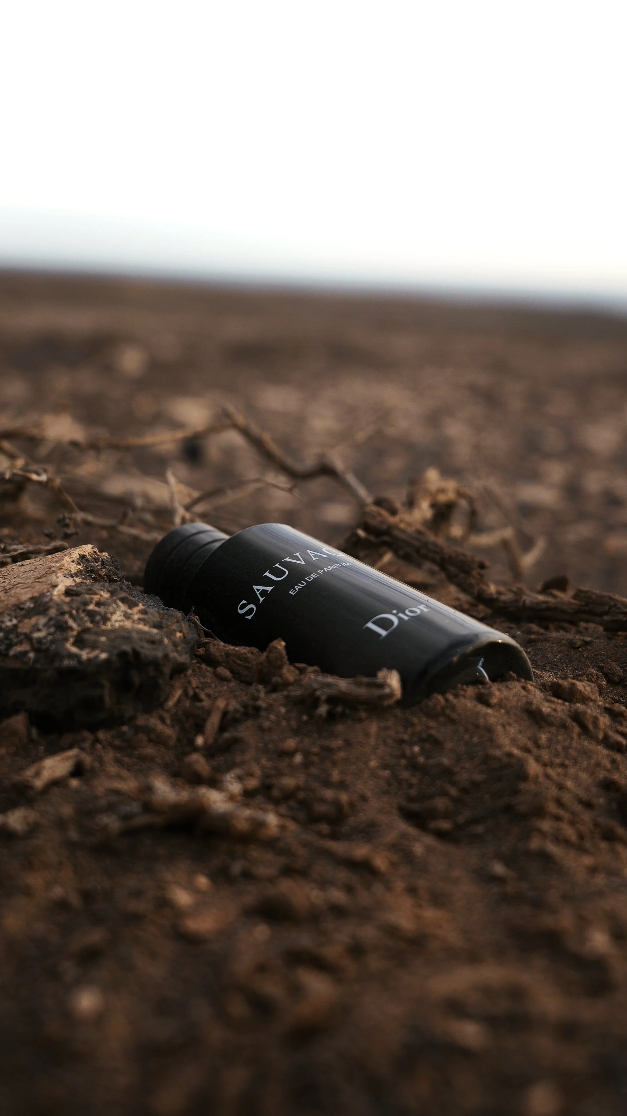A black bottle of Dior Sauvage perfume lying on brown dirt with twigs and debris, with a blurred horizon in the background.