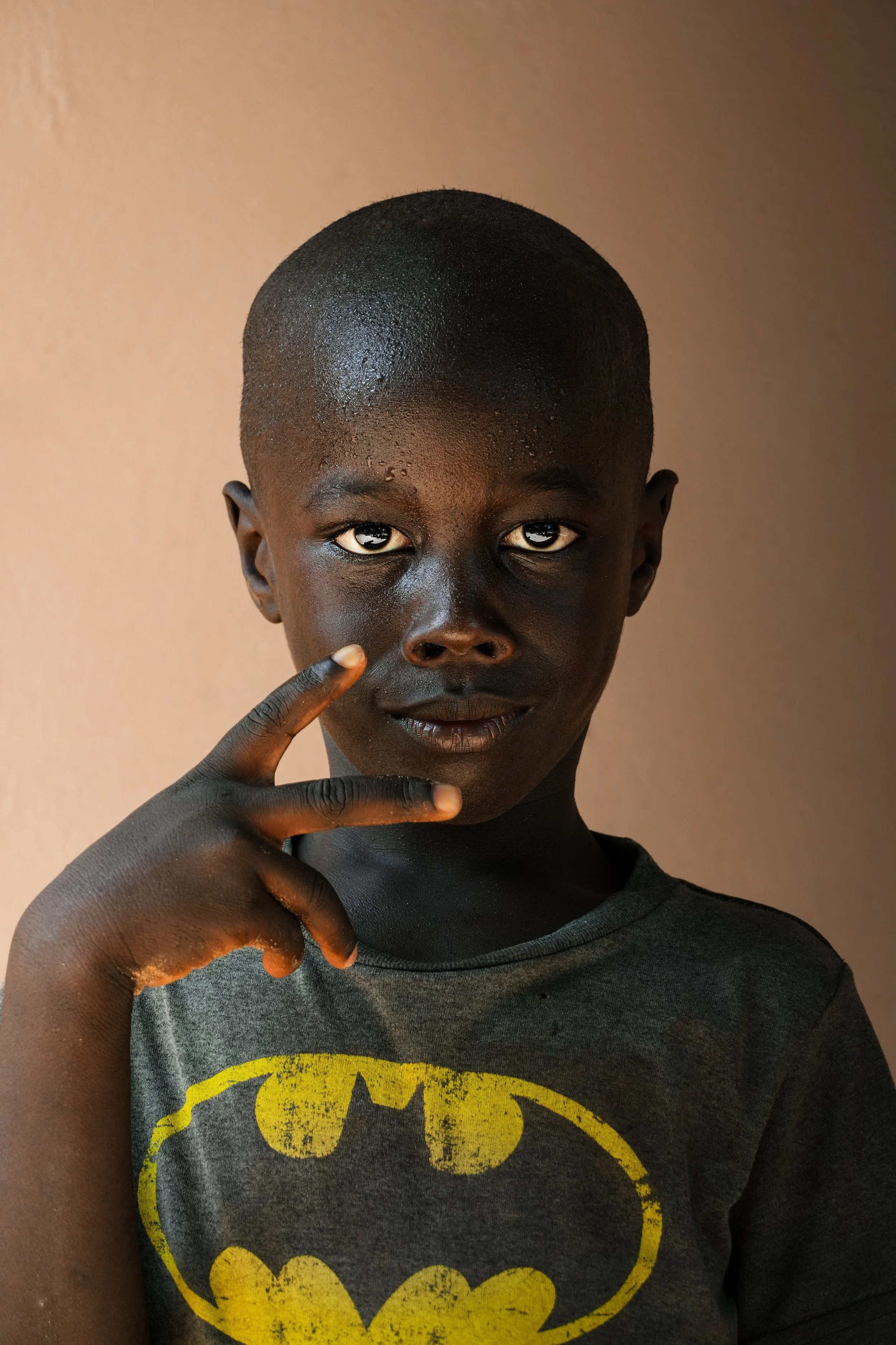 A young boy with dark skin and a close-cropped haircut posing indoors against a plain background, wearing a gray T-shirt with a yellow Batman logo, and making a peace sign with his hand near his face.