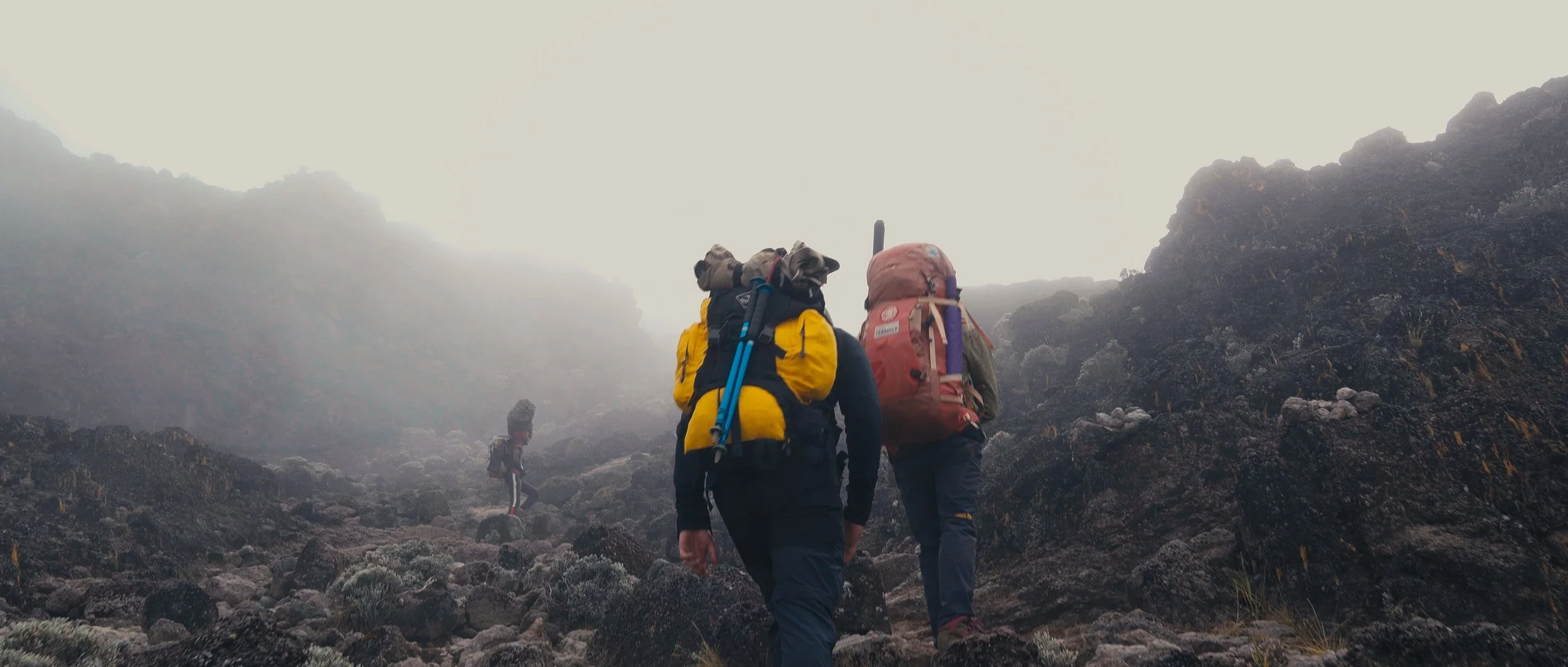 Three hikers with backpacks trekking up a foggy, rocky mountain trail.