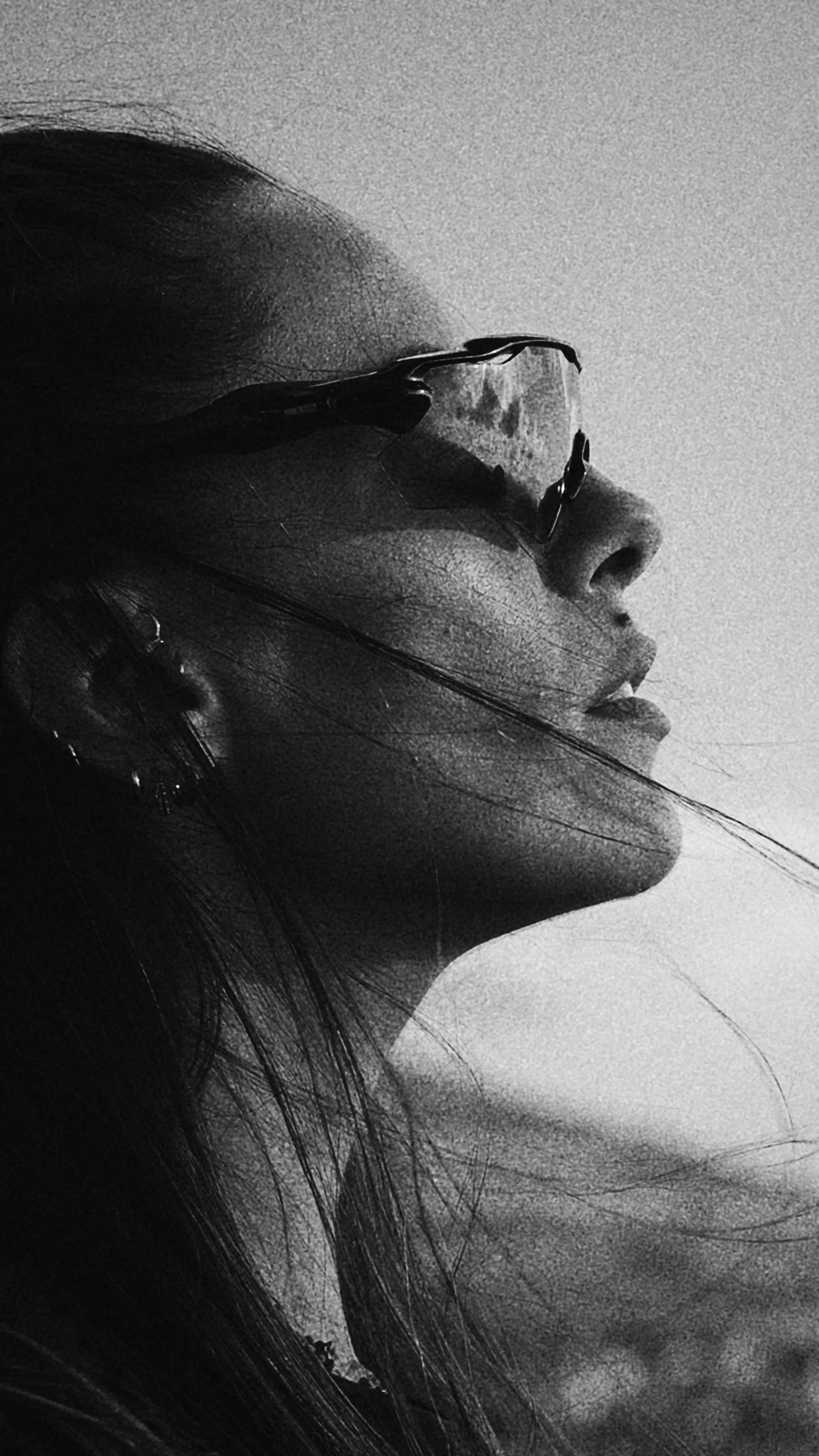 Close-up black and white photo of a woman's face in profile, wearing sunglasses, with long hair flowing.
