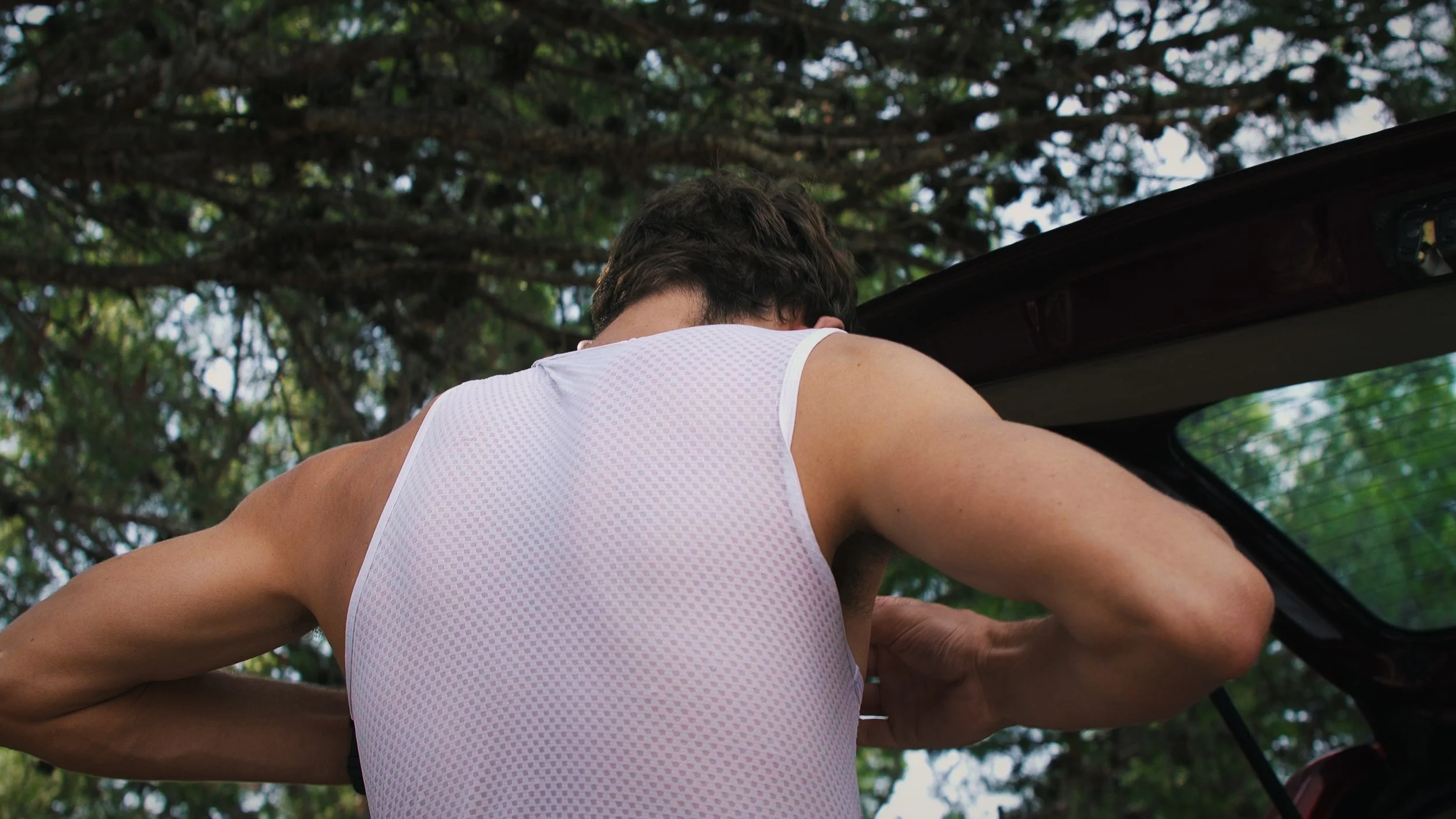 A man wearing a white sports tank top loading items into a car trunk outdoors, with trees in the background.
