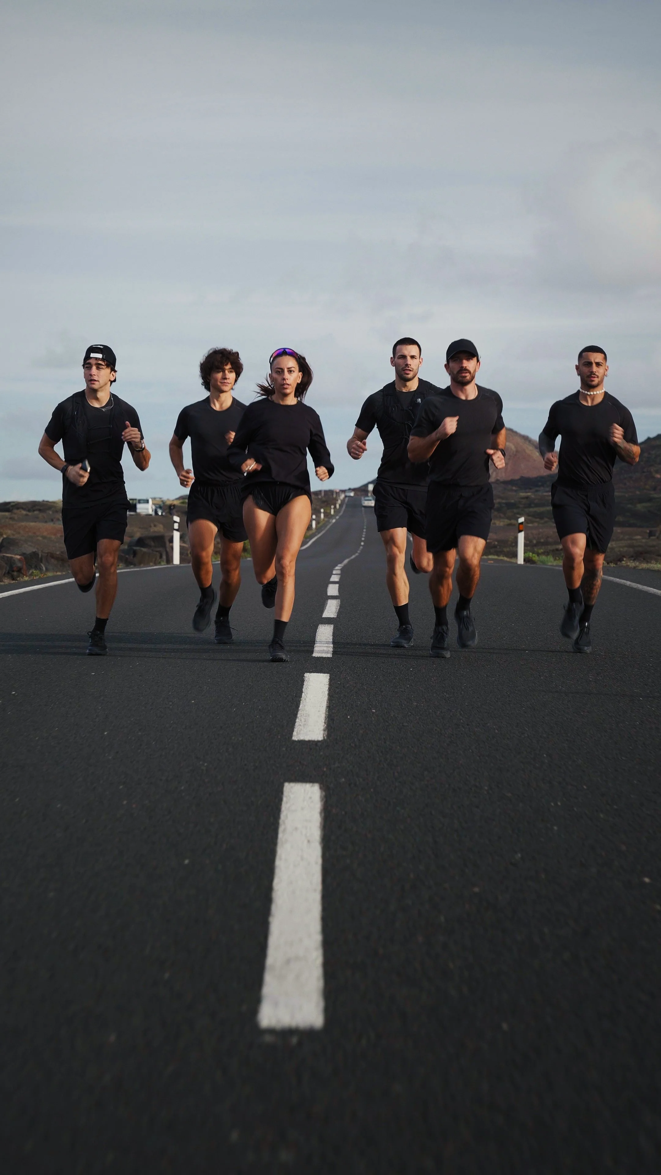 Six runners jogging on an open road in a rural landscape, wearing black athletic clothing.