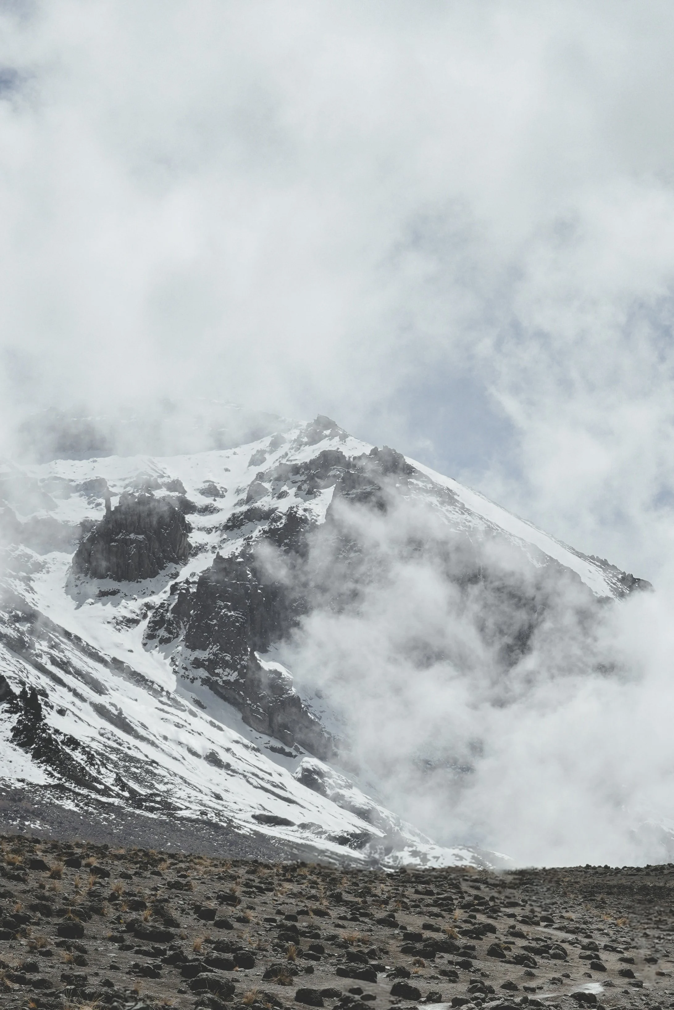 Snow-covered mountain peak shrouded in clouds, with rocky terrain in the foreground.