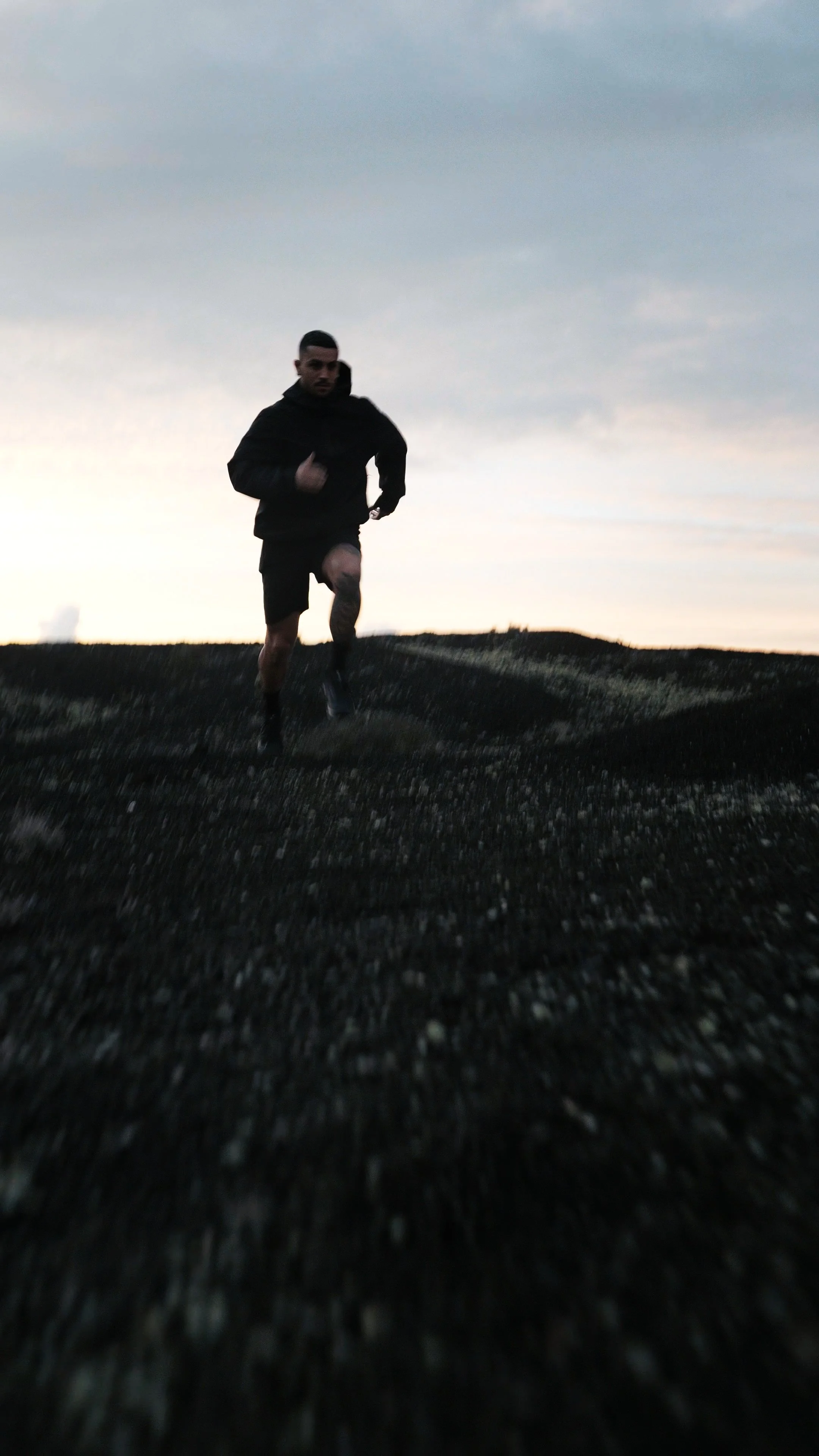 A man running on a dark, textured uneven terrain at dusk with a cloudy sky in the background.