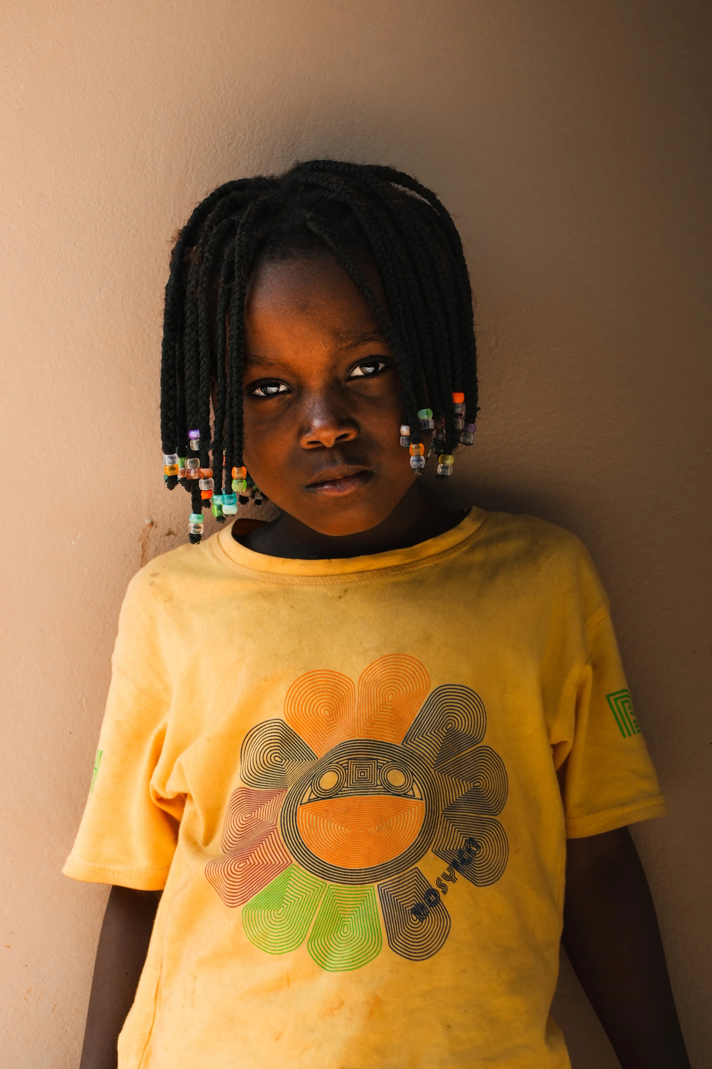 A young girl with braided hair decorated with colorful beads, leaning against a beige wall, wearing a yellow T-shirt with a multicolored circular design and text.