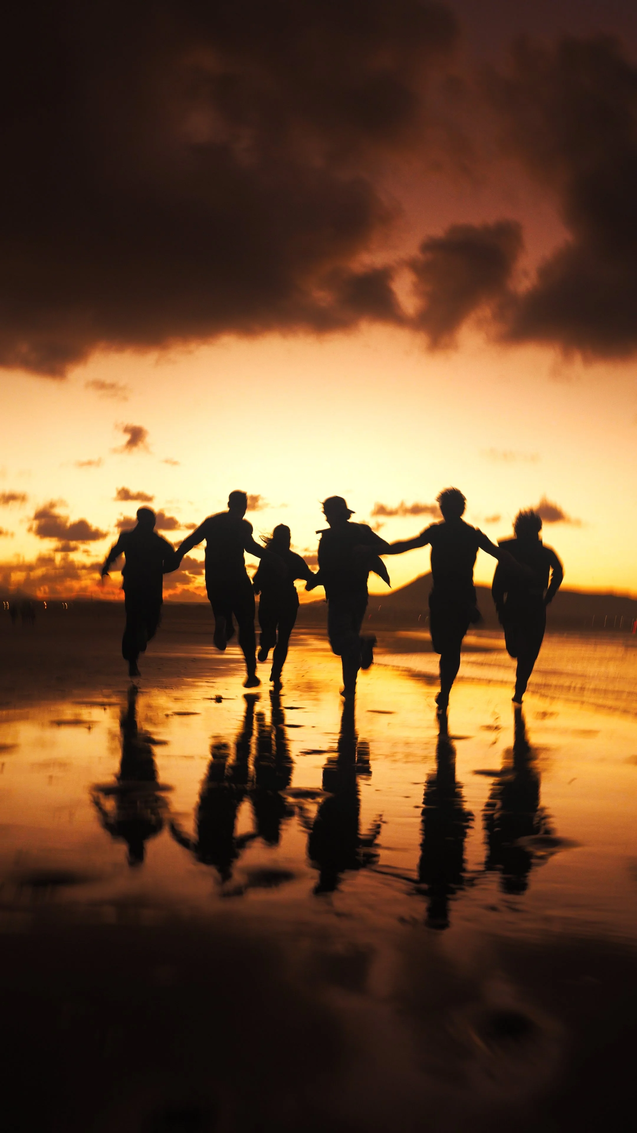 Silhouettes of seven people running on the beach at sunset, with clouds in the sky and their reflections visible on the wet sand.