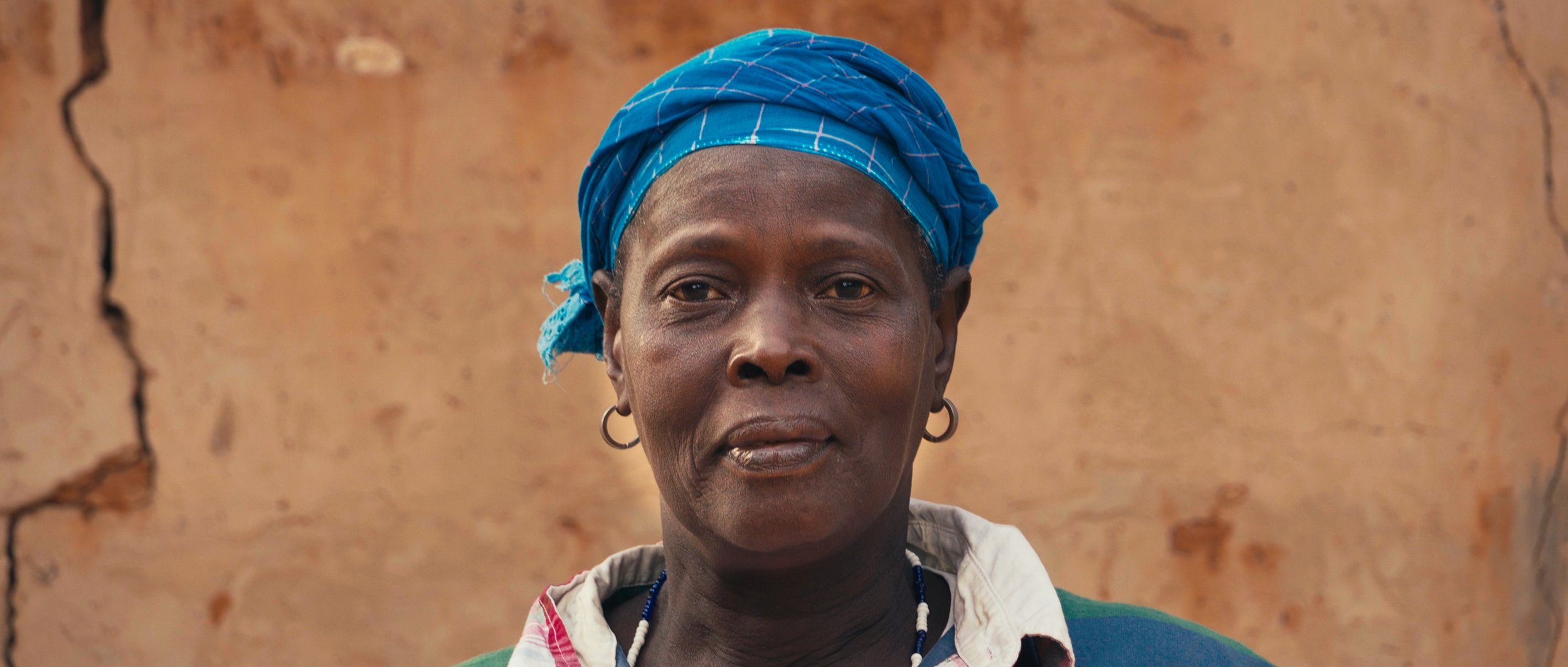 Close-up of a woman wearing a blue headscarf, earrings, and a necklace, standing outdoors with a cracked brown wall in the background.