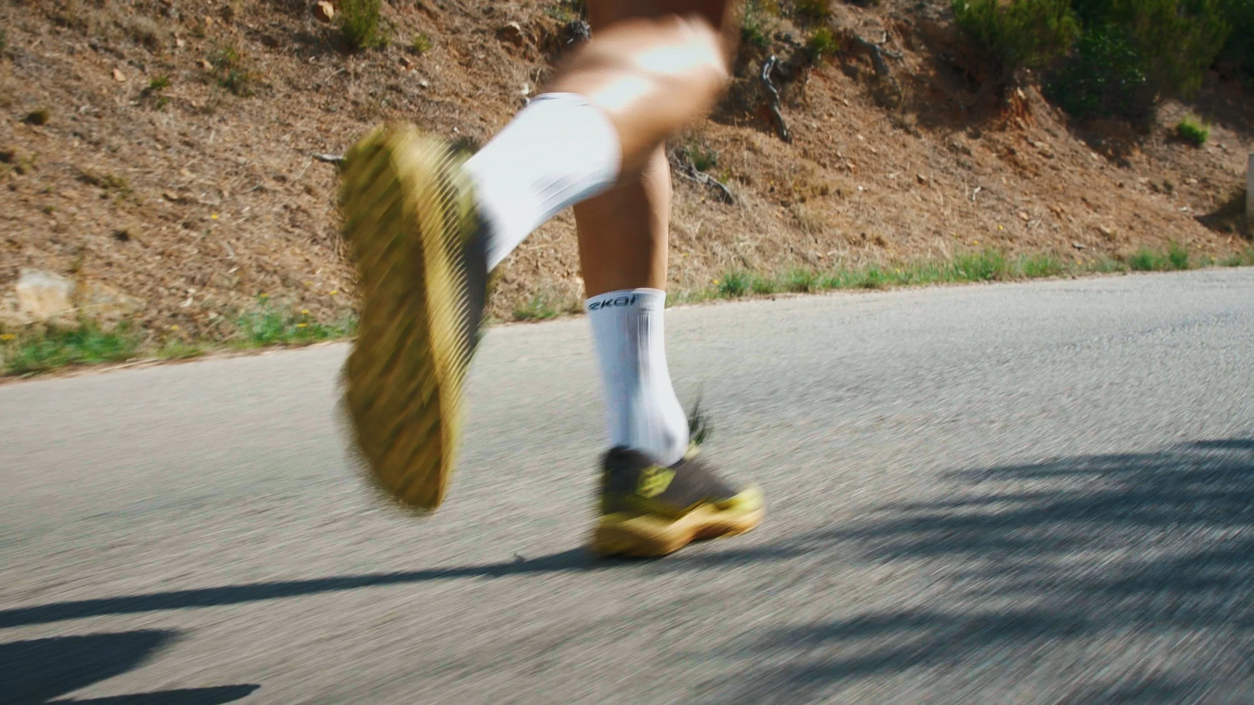 Close-up of a person running on a paved road, blurred motion, wearing white shorts, black running shoes with yellow accents, and white socks, with a dirt and grassy hillside on the side.