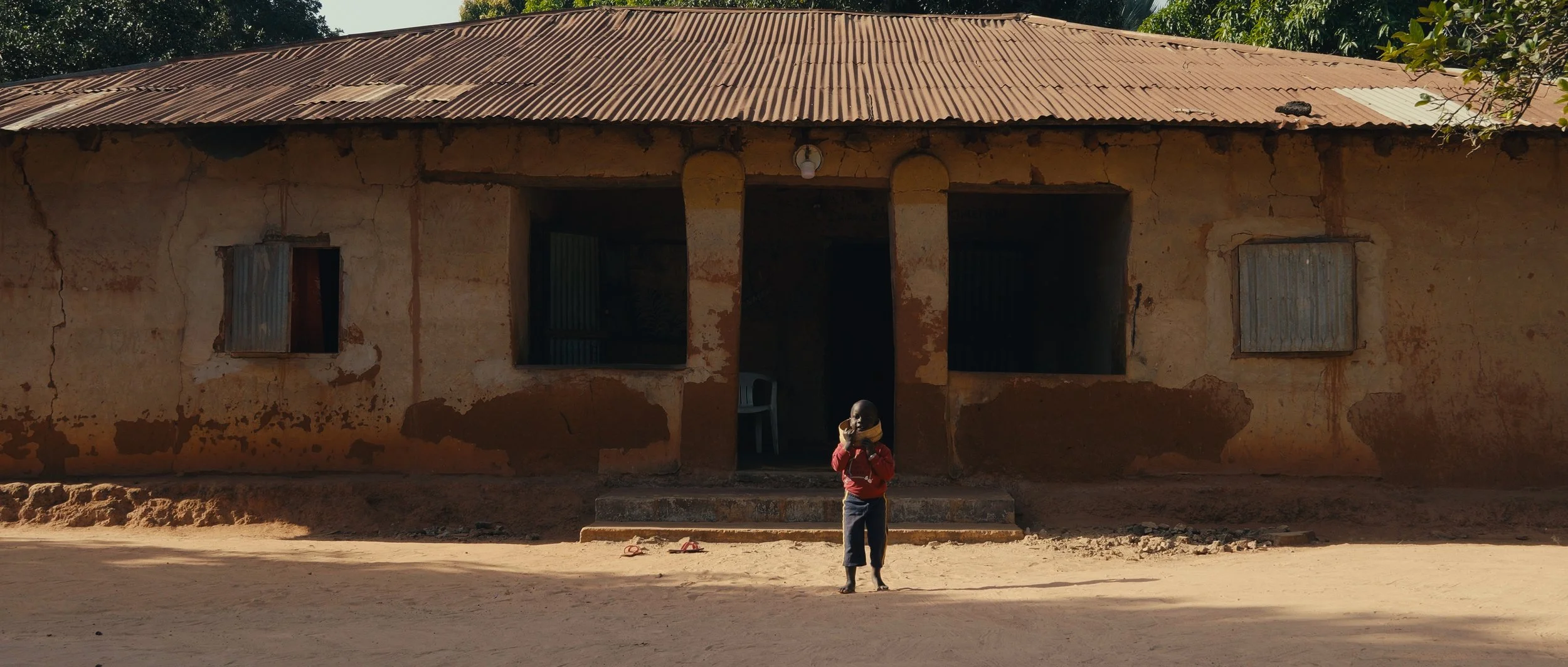 A young child standing in front of an old, weathered house with a metal roof, holding a phone to their ear.