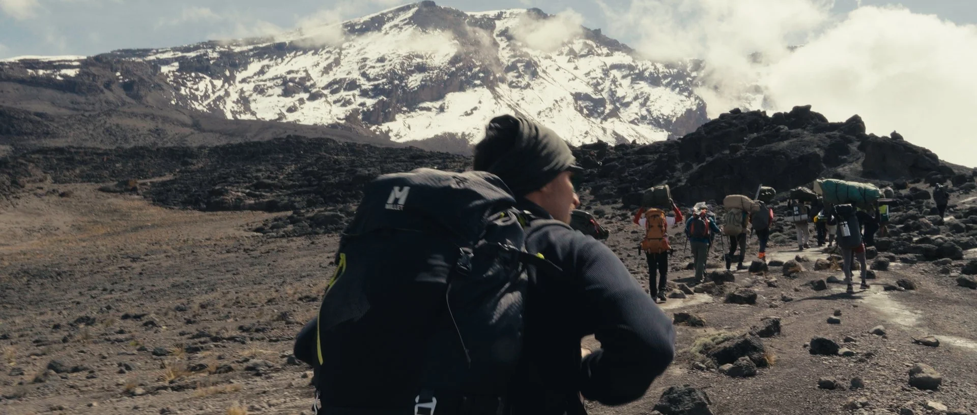 Group of hikers walking on a rocky mountain trail with snow-capped peaks in the background.