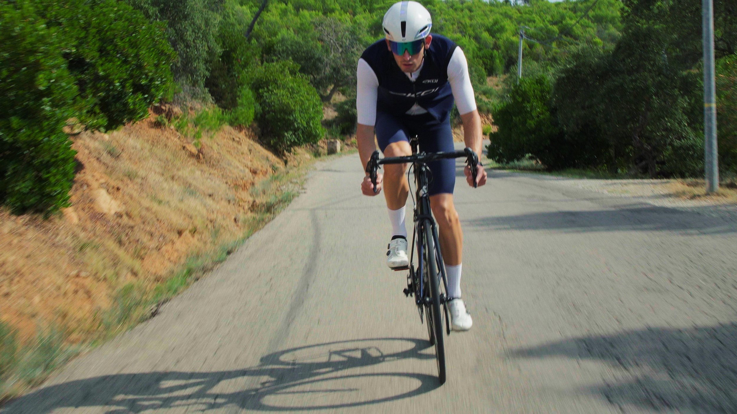 A man riding a black road bicycle on a winding paved road surrounded by greenery and trees, wearing a white helmet, sunglasses, a black and white sleeveless cycling jersey, navy cycling shorts, and white shoes.