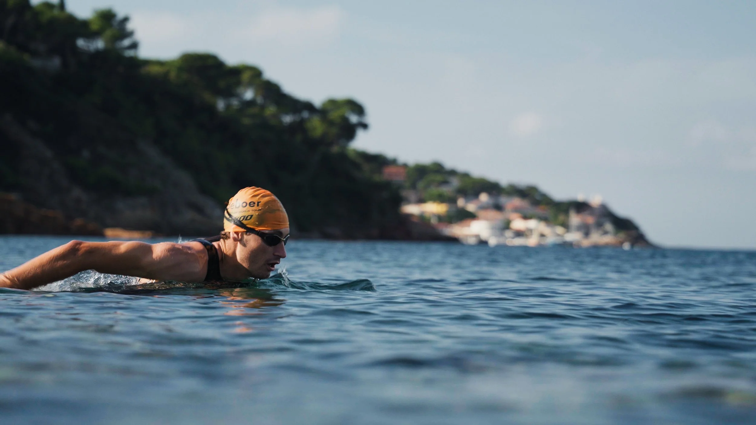 Swimmer in a black swimsuit with an orange swim cap and goggles, swimming in open water near a coastline with greenery and houses in the background.