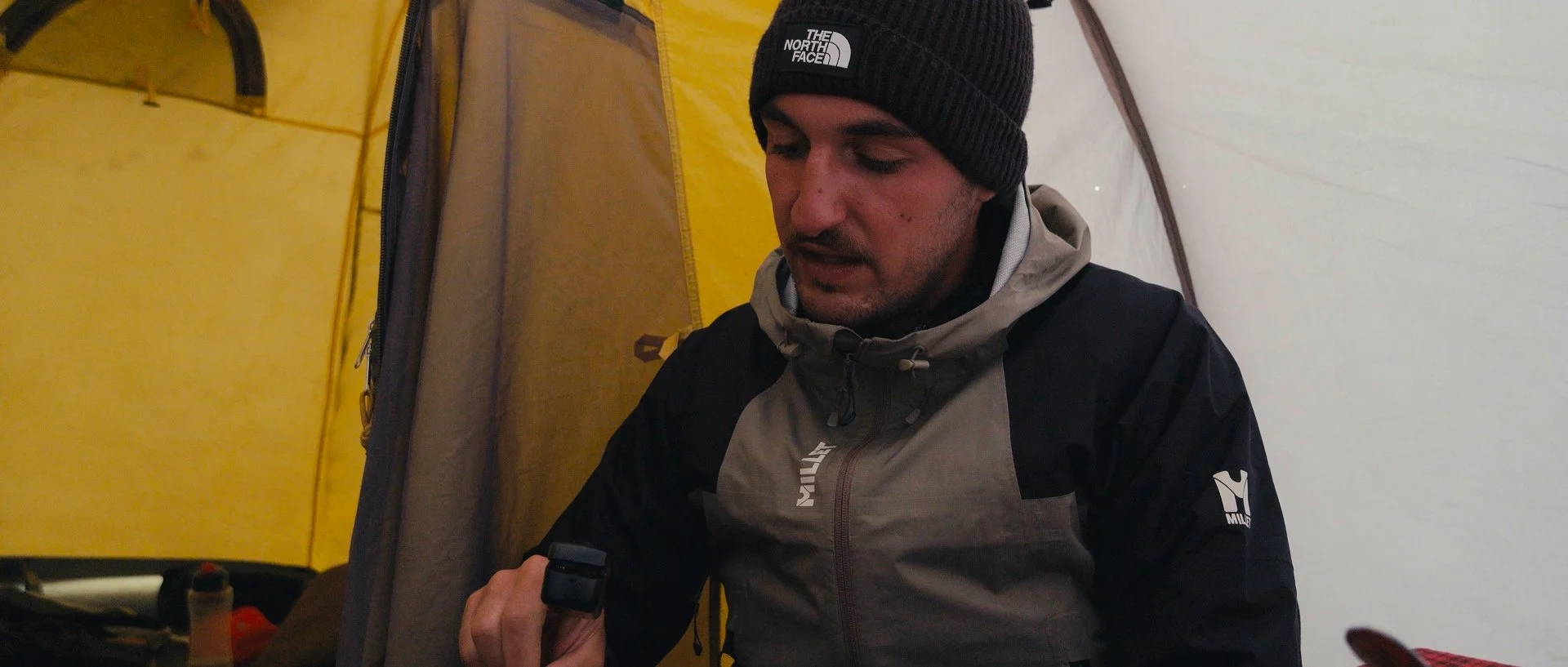 A man wearing a black beanie and a gray and black jacket inside a tent, looking at a device in his hand.