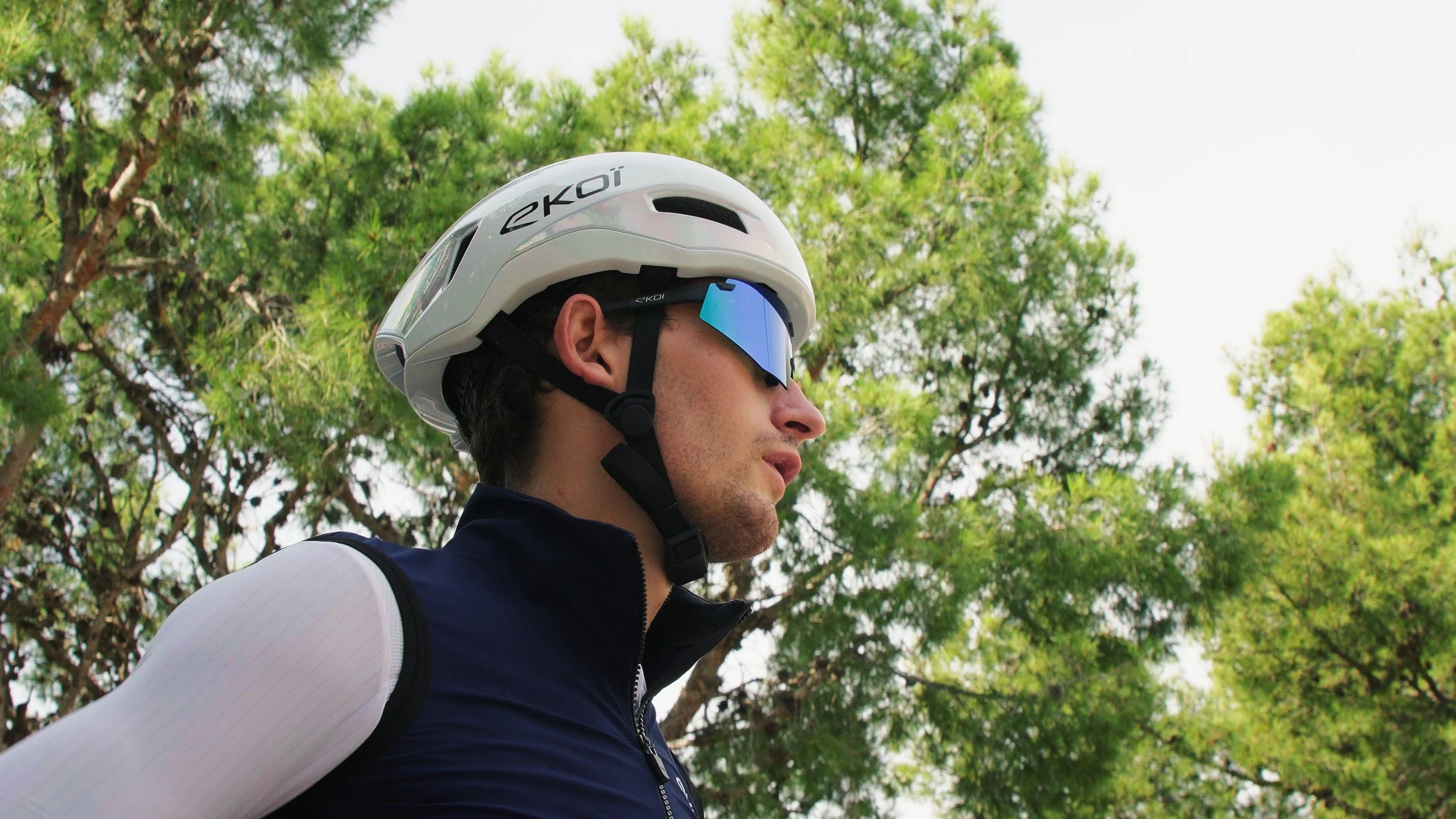 A man wearing a white bicycle helmet with the brand name 'ekoi,' reflective blue sunglasses, a navy blue vest, and a white long sleeve shirt underneath, stands outdoors with green trees in the background.