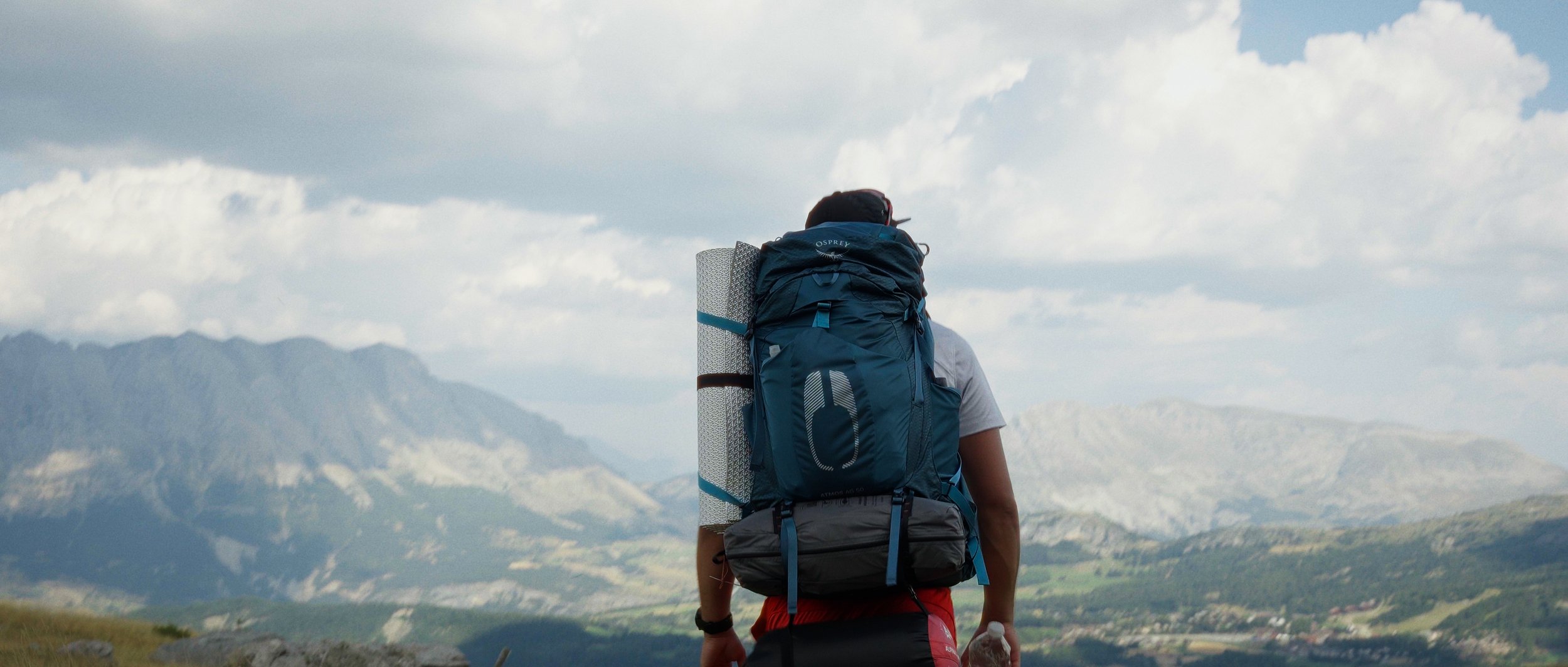 Backpacker carrying a large blue backpack with a rolled sleeping pad, walking outdoors in mountainous terrain under cloudy sky.