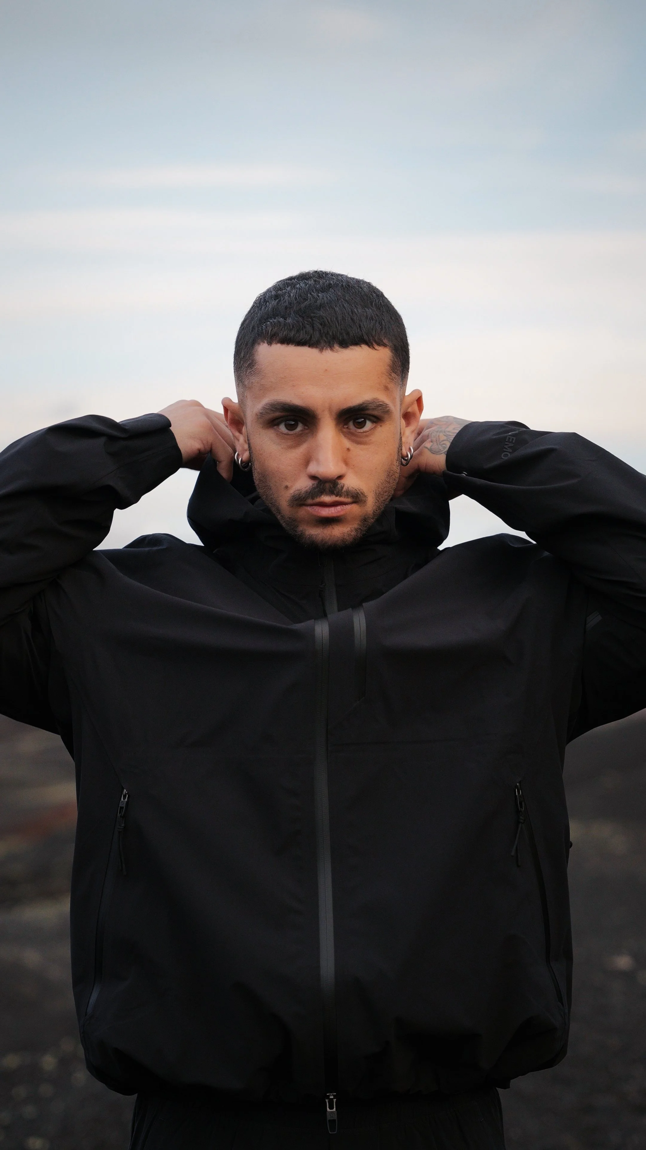 A man with short dark hair and earrings, wearing a black jacket, is standing outdoors with a cloudy sky in the background, looking directly at the camera and adjusting his jacket hood.