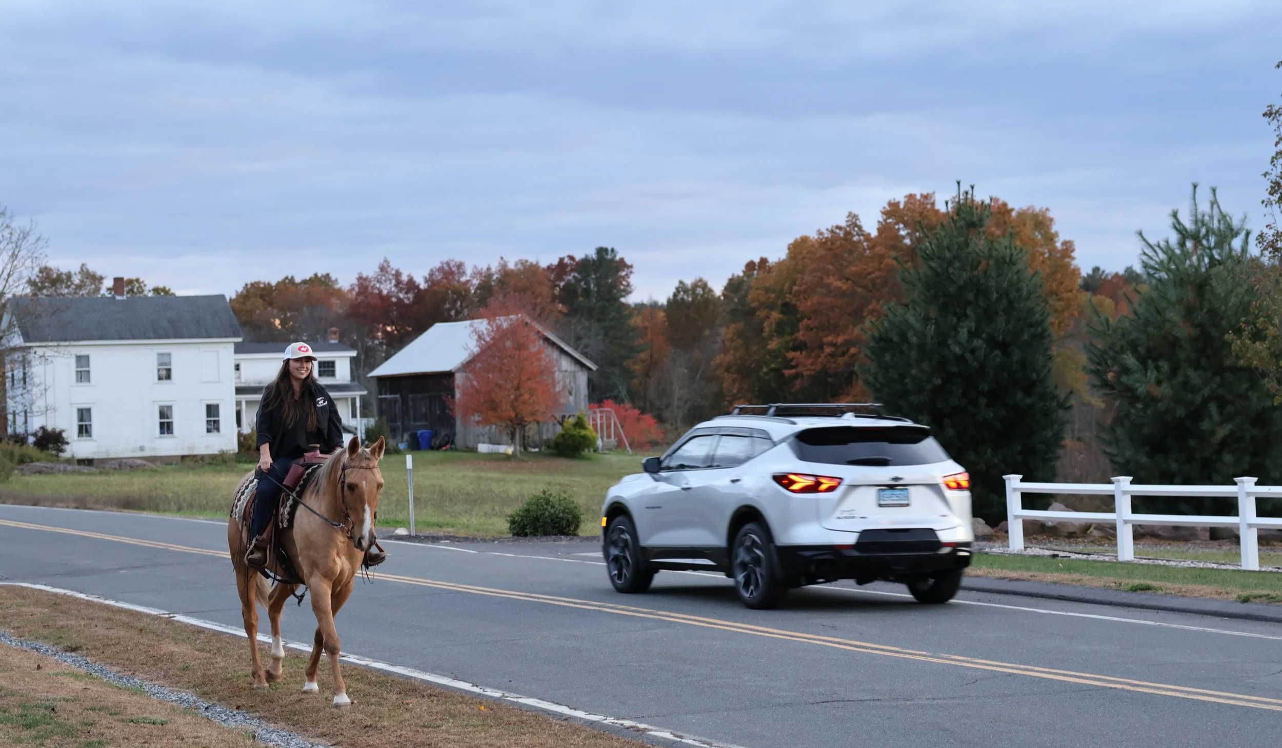 A woman riding a light brown horse along a suburban street with trees showing fall foliage, a white house, a barn, and a white SUV driving by.