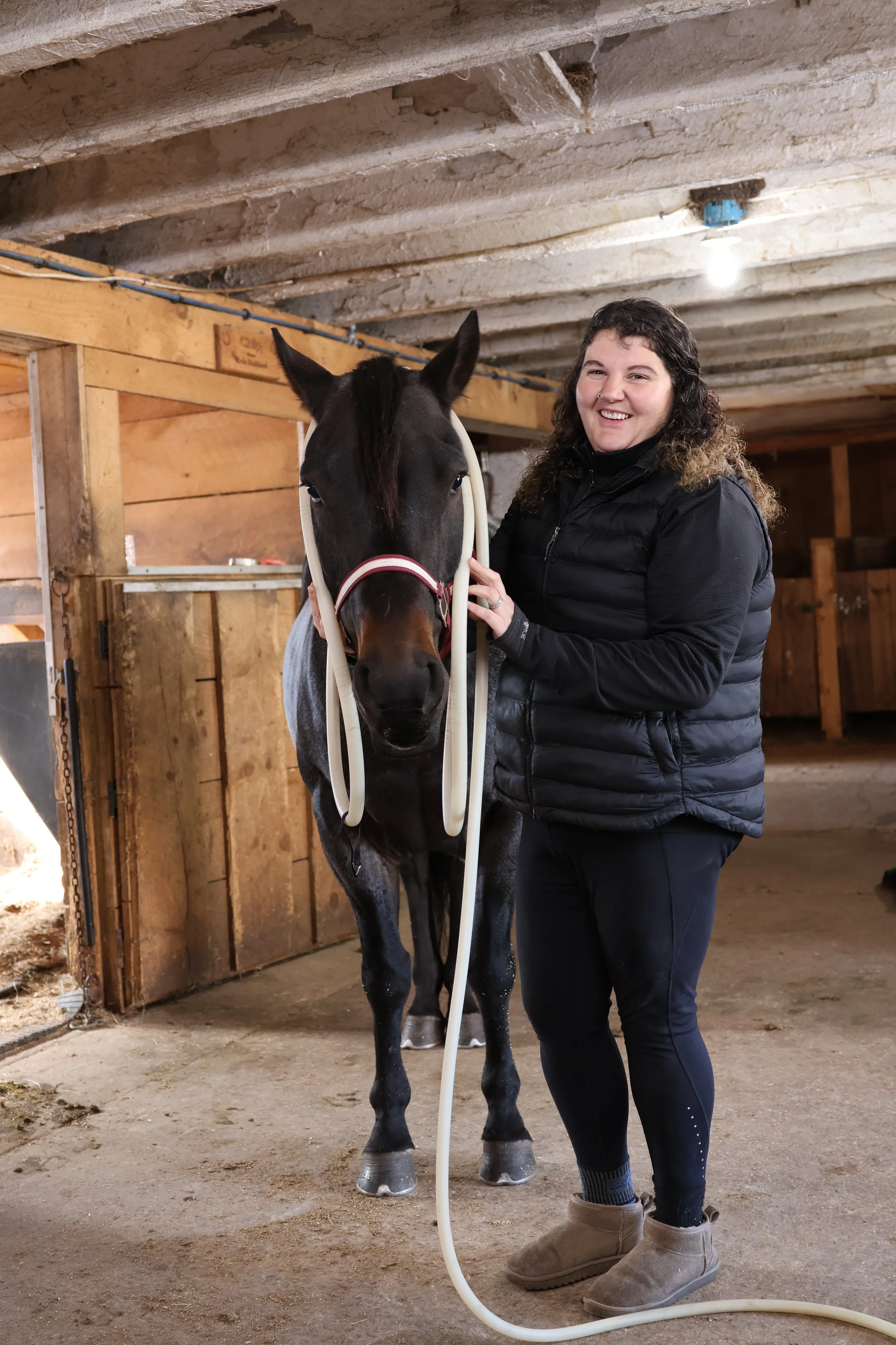 A woman standing with a black horse inside a stable, holding a hose near the horse's face, both smiling.