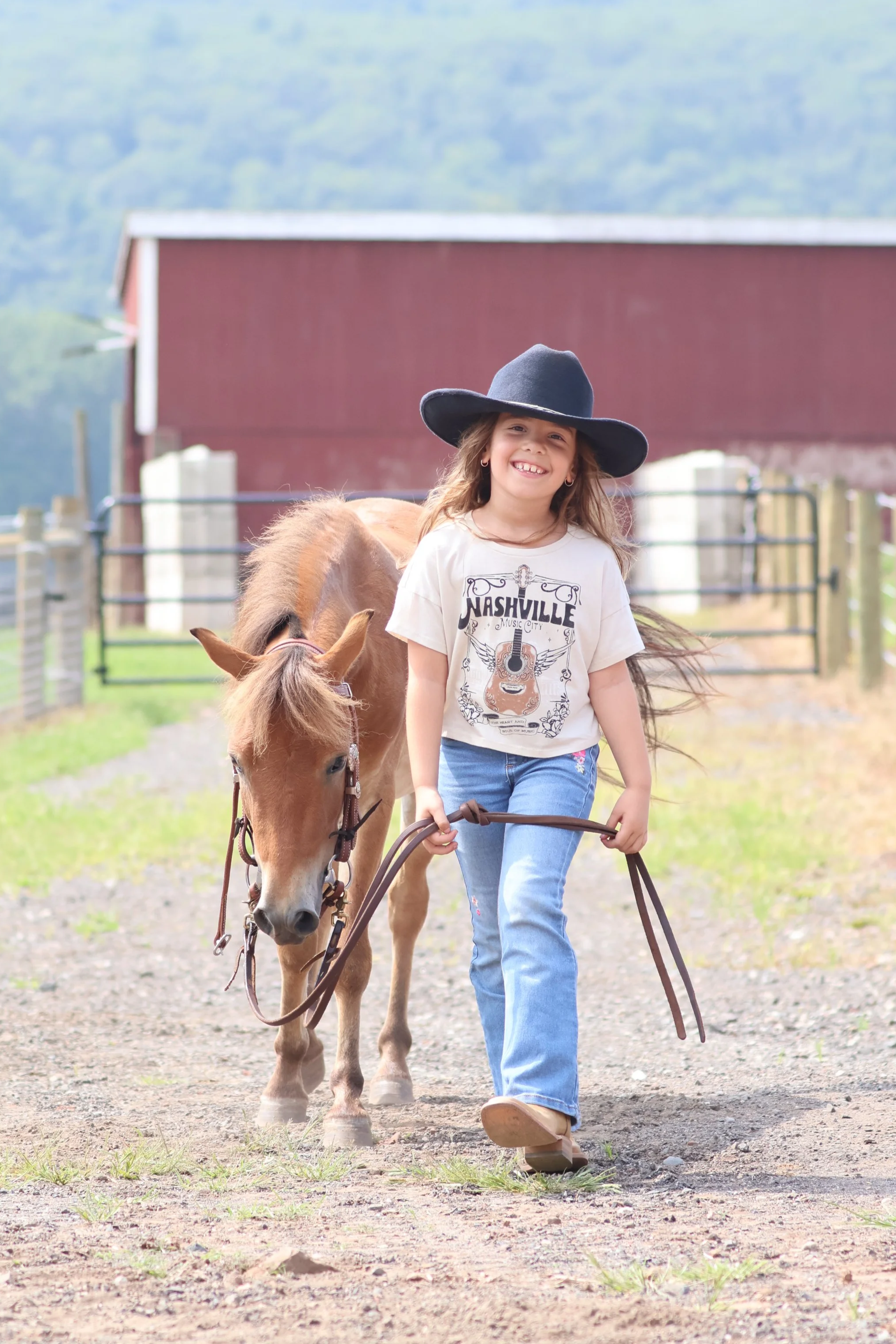 A young girl with long hair, wearing a wide-brimmed black hat, jeans, and a Nashville music T-shirt, walks a brown pony on a dirt path in a country setting with a red barn and green hills in the background, smiling.