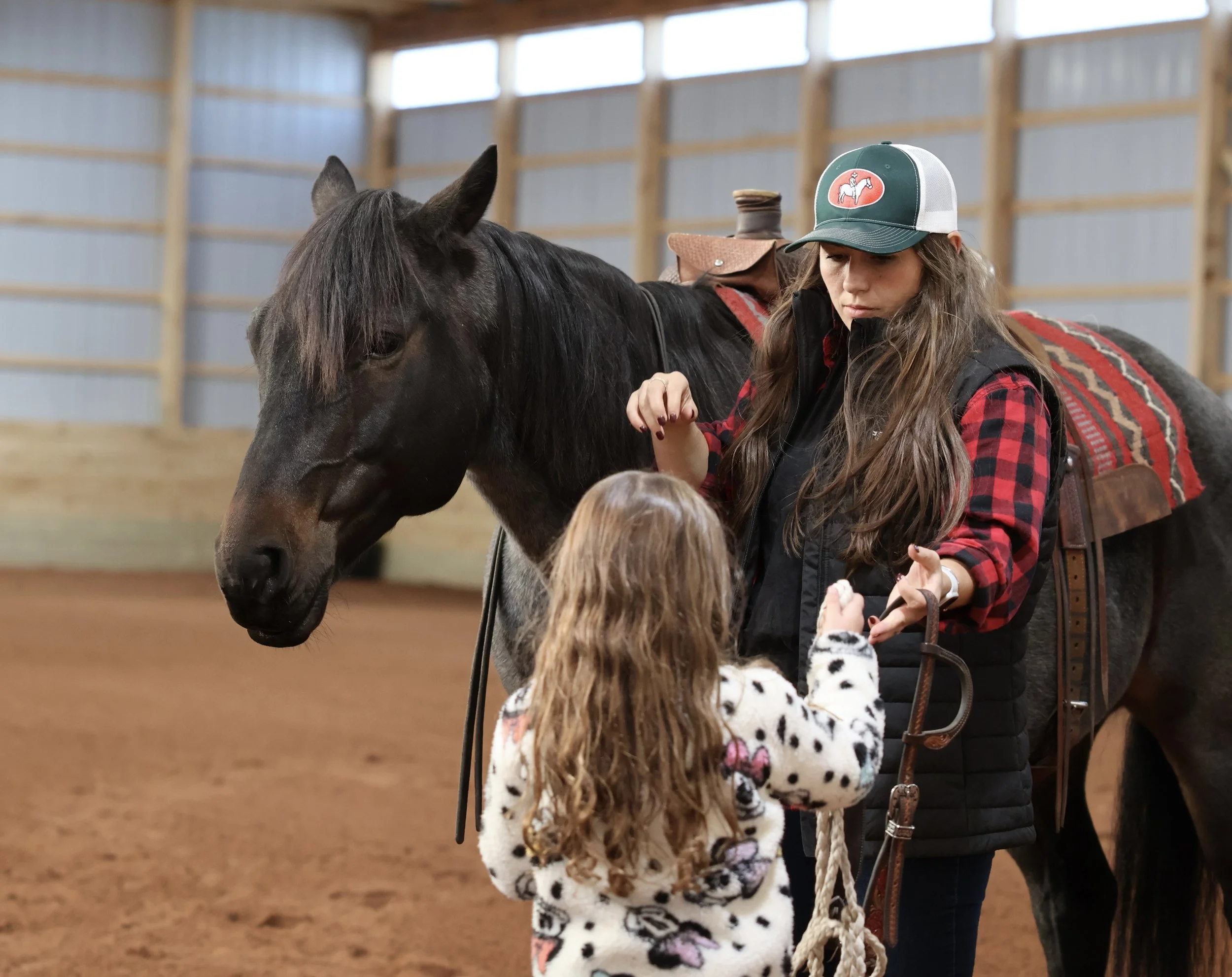 A woman and a young girl standing next to a dark horse inside an indoor riding arena. The woman is wearing a cap, a red and black plaid shirt under a black vest, and the girl is wearing a spotted jacket. The woman appears to be preparing the horse for riding.