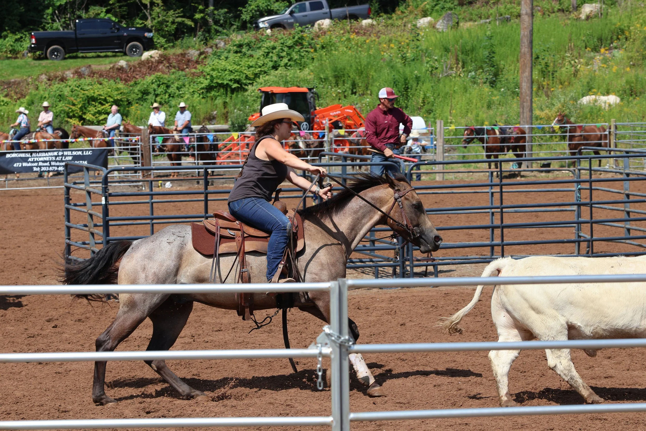 People riding horses in an outdoor arena with a dirt ground, fenced with metal railings, and a green, hilly background with vehicles and trees.