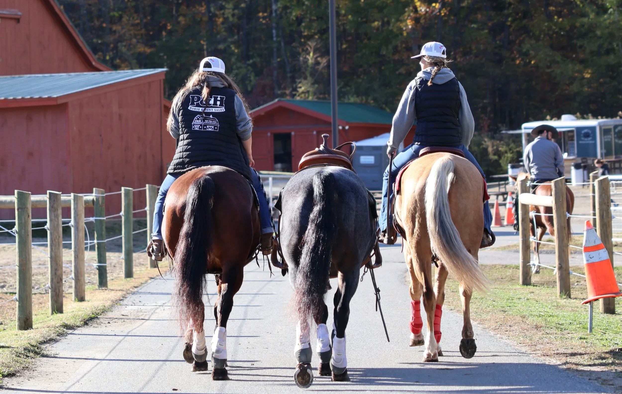 Three people riding horses on a trail near a farm or ranch. The riders are wearing casual riding attire, including baseball caps and vests. The horses are walking on a paved pathway surrounded by wooden fencing, with red barns and other farm structures in the background.