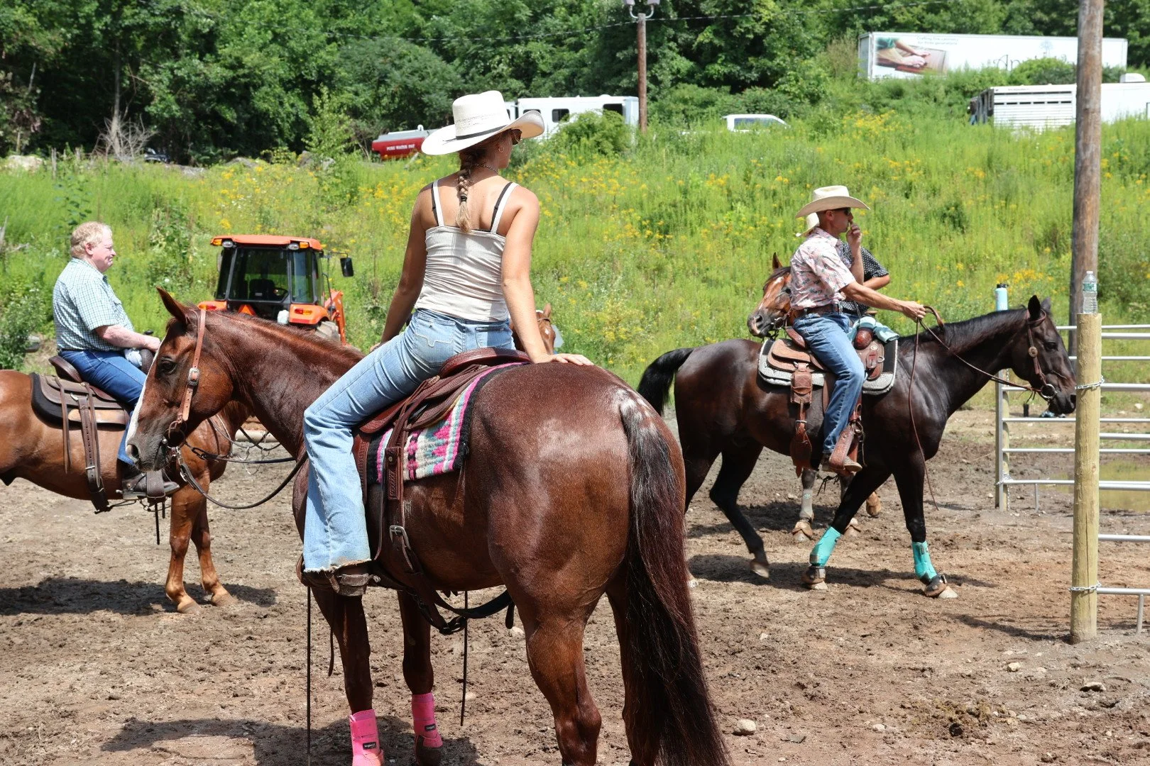 Three people riding horses on a dirt trail in a rural setting, with greenery and trees in the background.