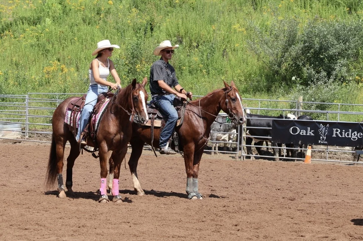 Two people riding horses in an outdoor arena, wearing cowboy hats, with horses wearing leg wraps, and a sign that reads 'Oak Ridge Farm' in the background.