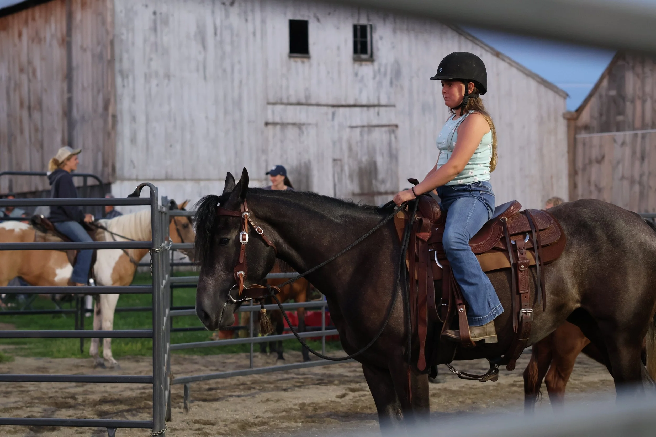 A young girl wearing a black riding helmet and sleeveless top riding a dark brown horse in an outdoor riding arena with a rustic barn in the background.