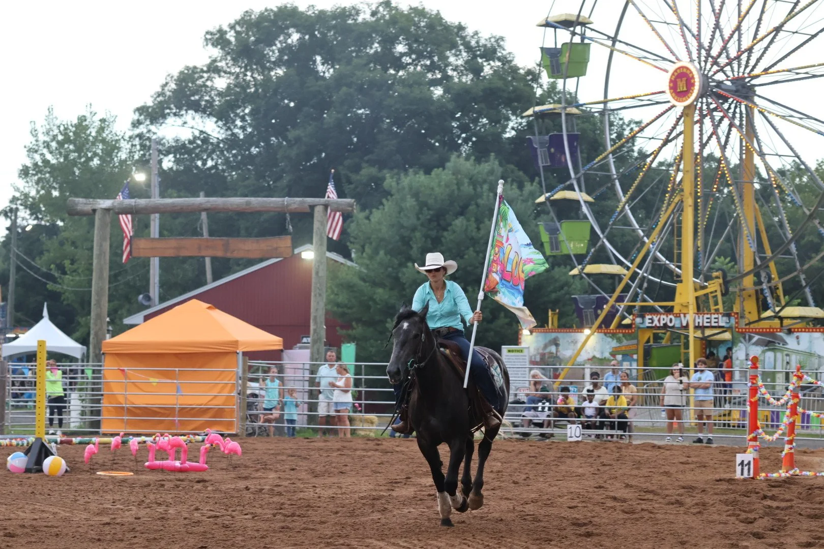 A woman riding a horse at a fair, holding a colorful flag. In the background, there is a ferris wheel, carnival tents, and a crowd of people watching.