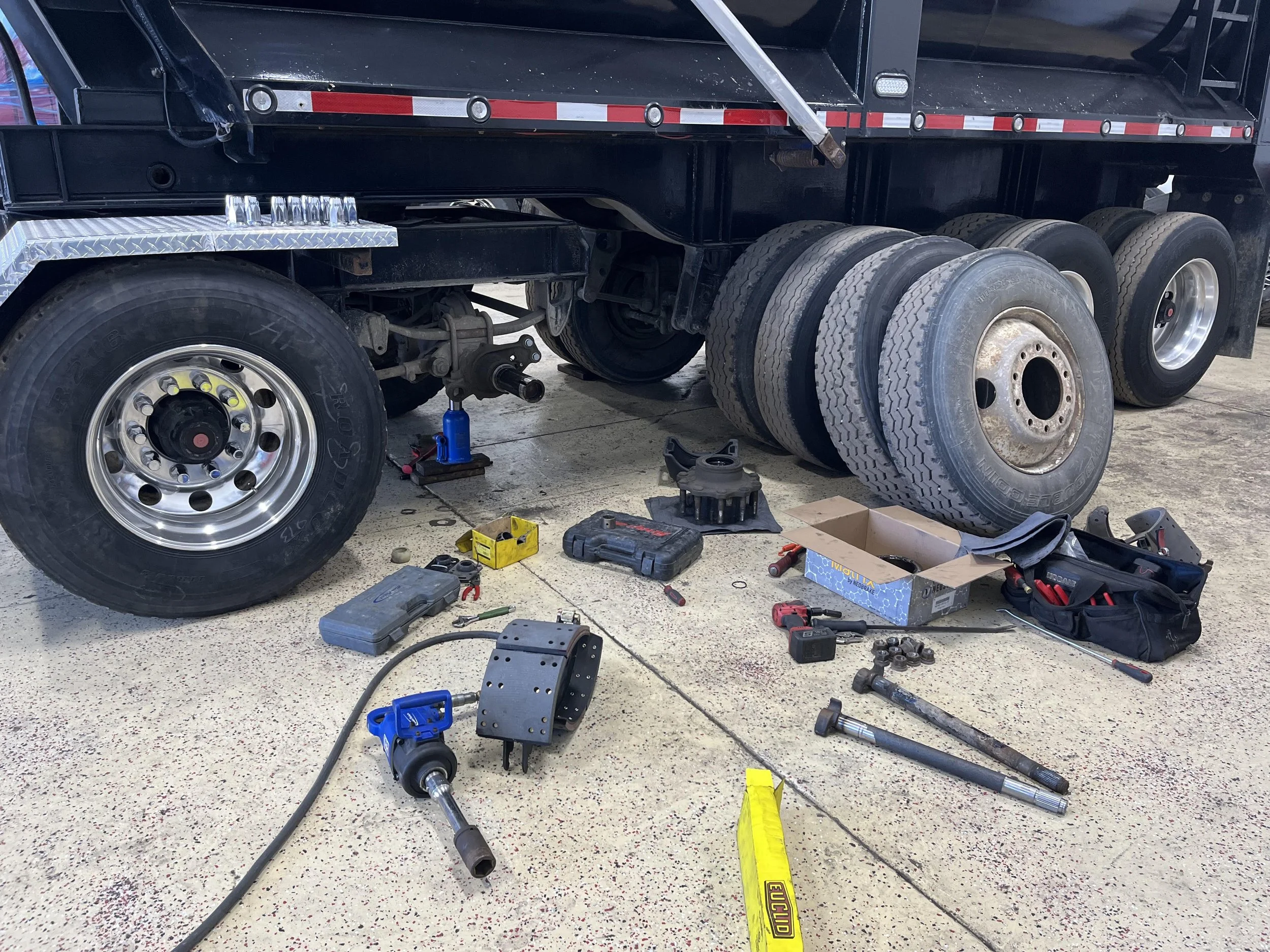 Rear view of a semi-truck with the wheels removed, showing tools and spare tires on the ground, as the truck is being repaired or maintained.