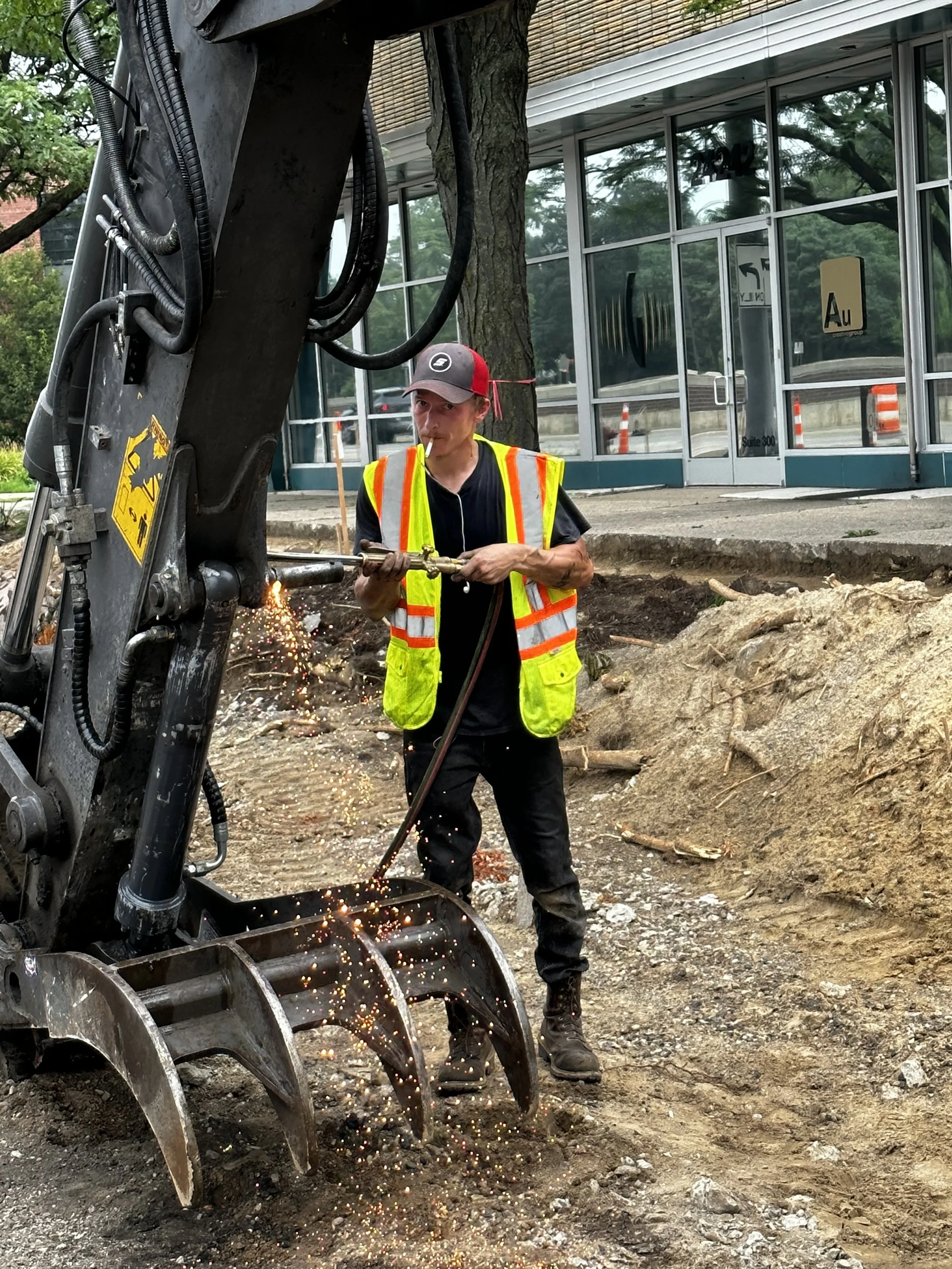 A construction worker wearing a yellow safety vest and black pants operating an excavator attachment with sparks flying while working on a dirt ground outside a building with glass windows.