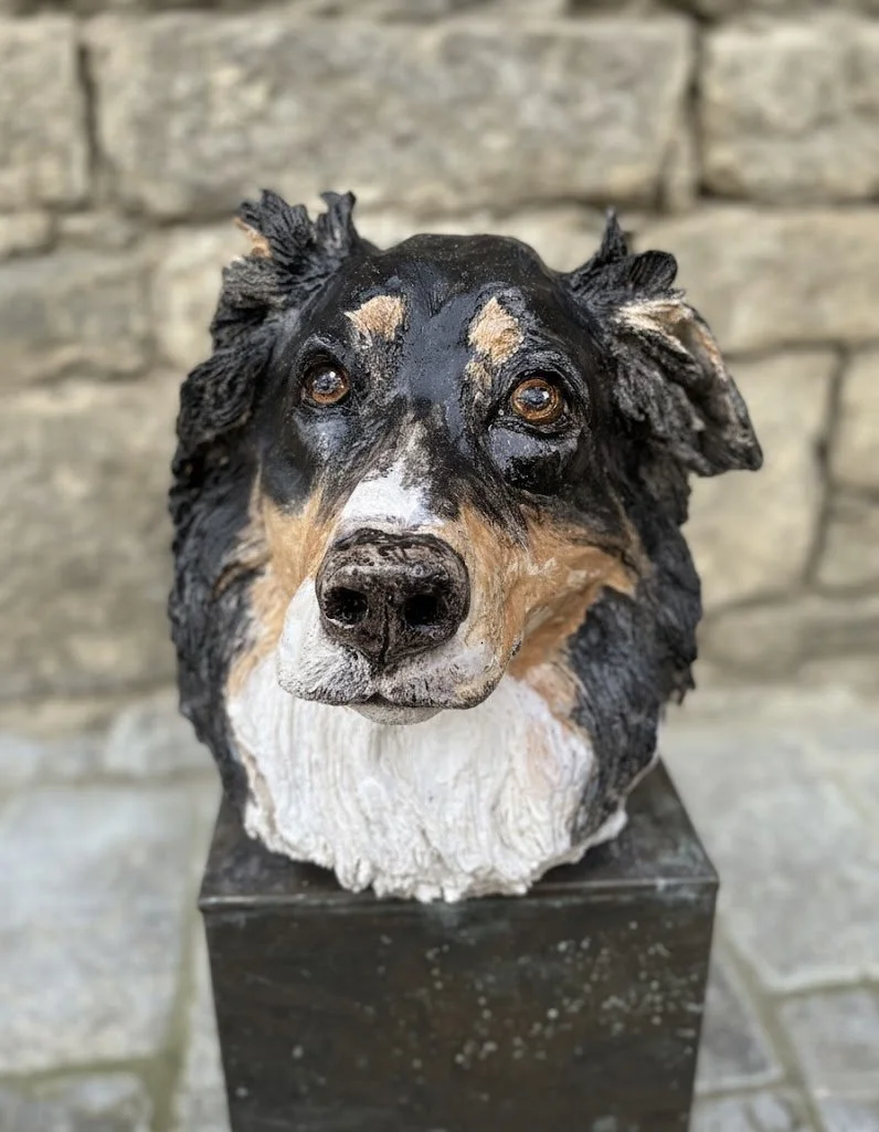A realistic sculpture of a dog, possibly an Australian Shepherd, with black, white, and tan fur, placed on a pedestal against a stone wall background.