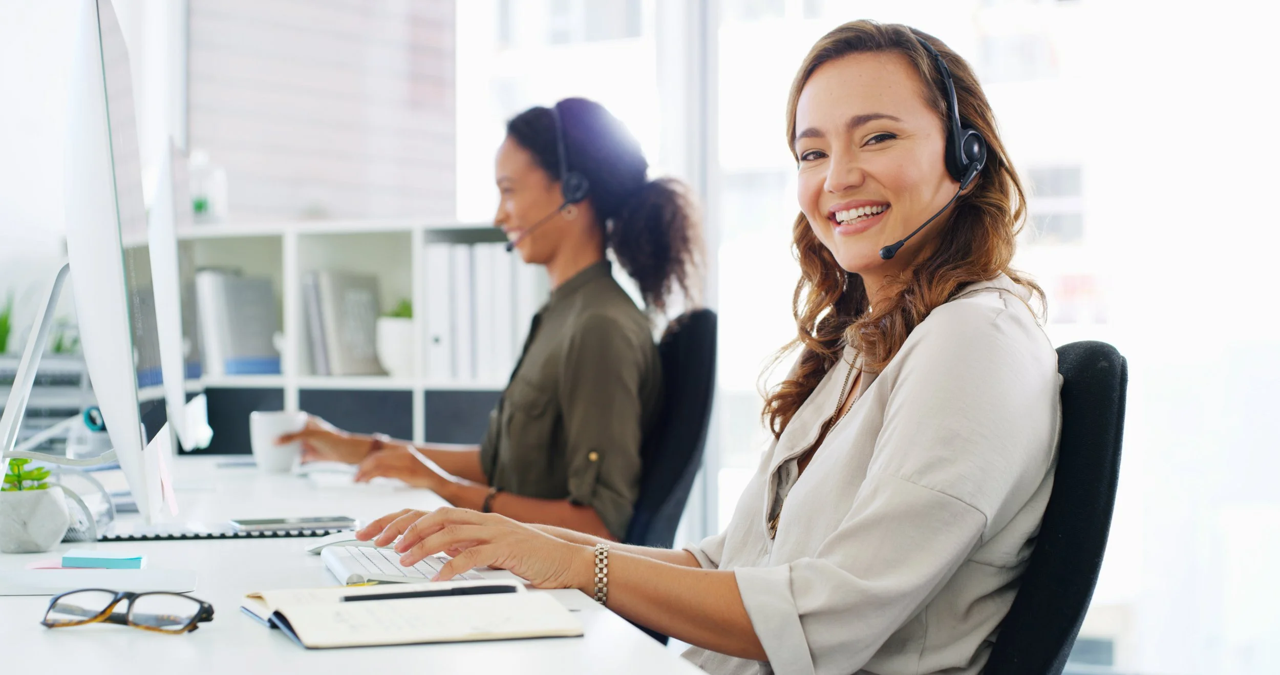 Two women working at computers in an office, both wearing headsets, with the woman in the foreground smiling and looking at the camera.