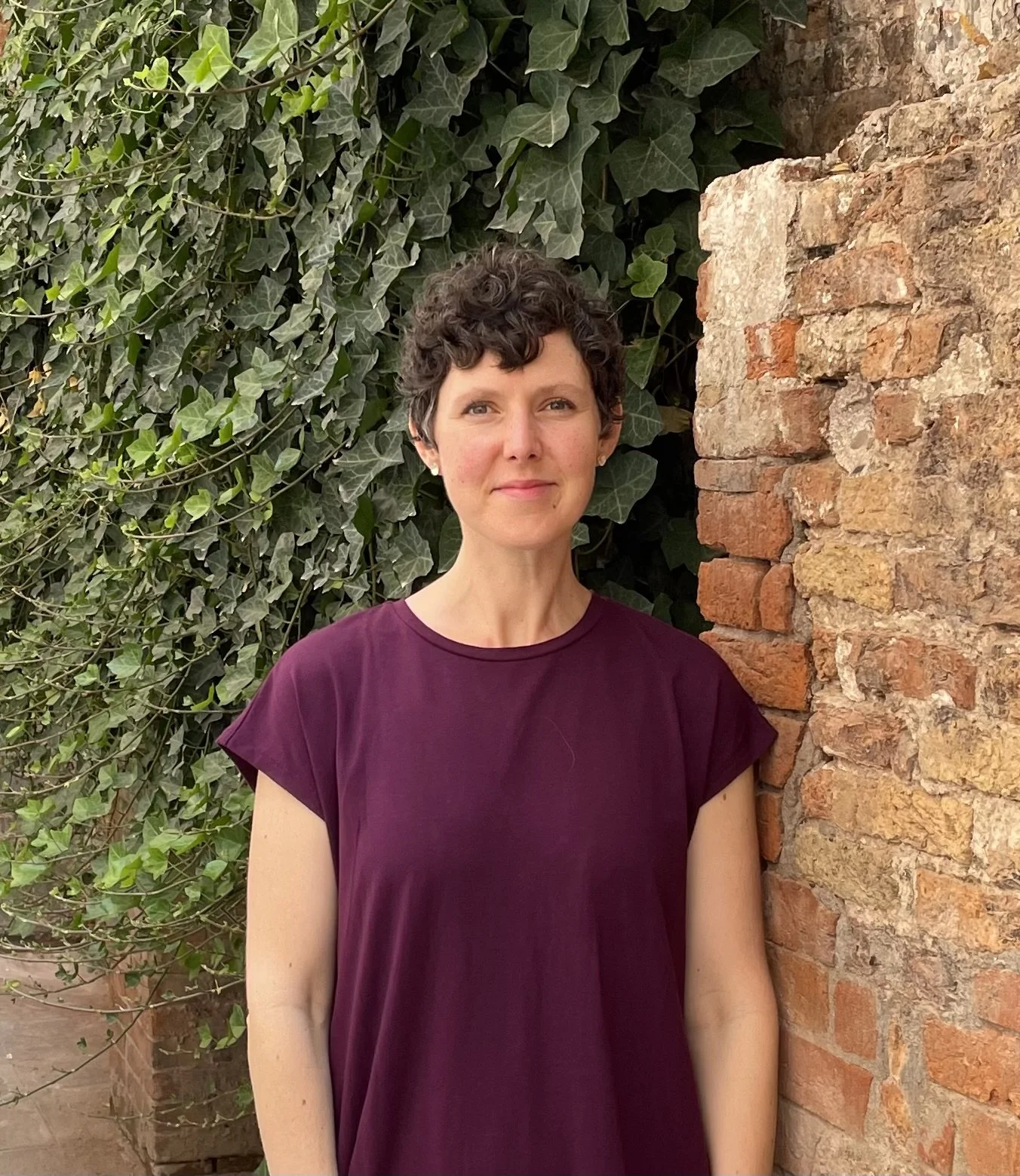 A woman with short curly hair wearing a purple shirt standing outdoors next to a brick wall, with green ivy leaves behind her.